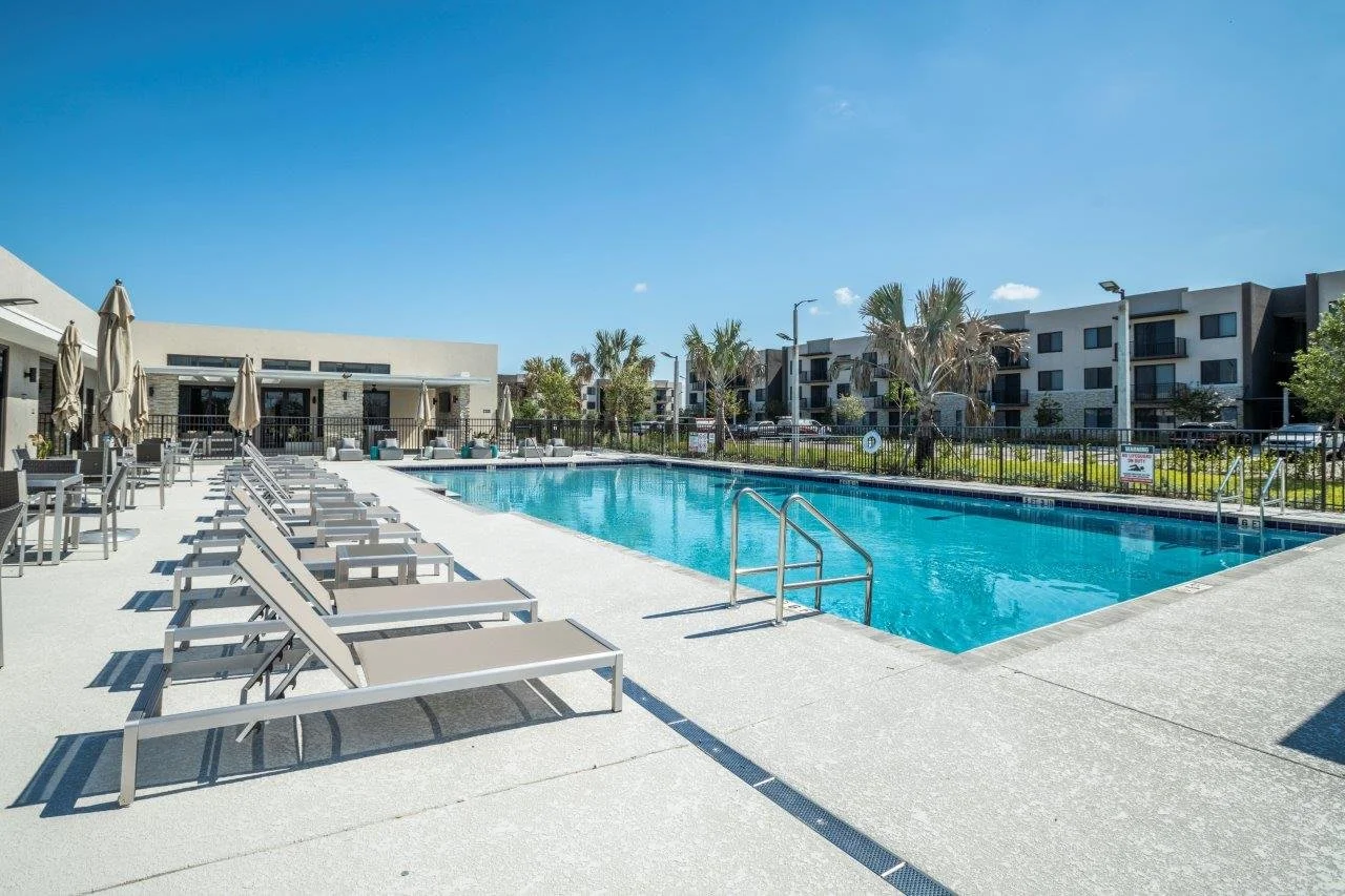 Empty outdoor swimming pool with lounge chairs and umbrellas, surrounded by apartment buildings and palm trees on a sunny day.