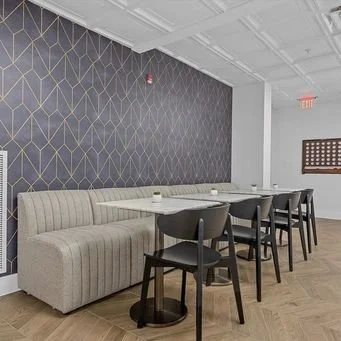 Interior of a cafe or restaurant with a long beige upholstered bench, four black chairs around a table, patterned wallpaper, and a white ceiling with decorative panels.