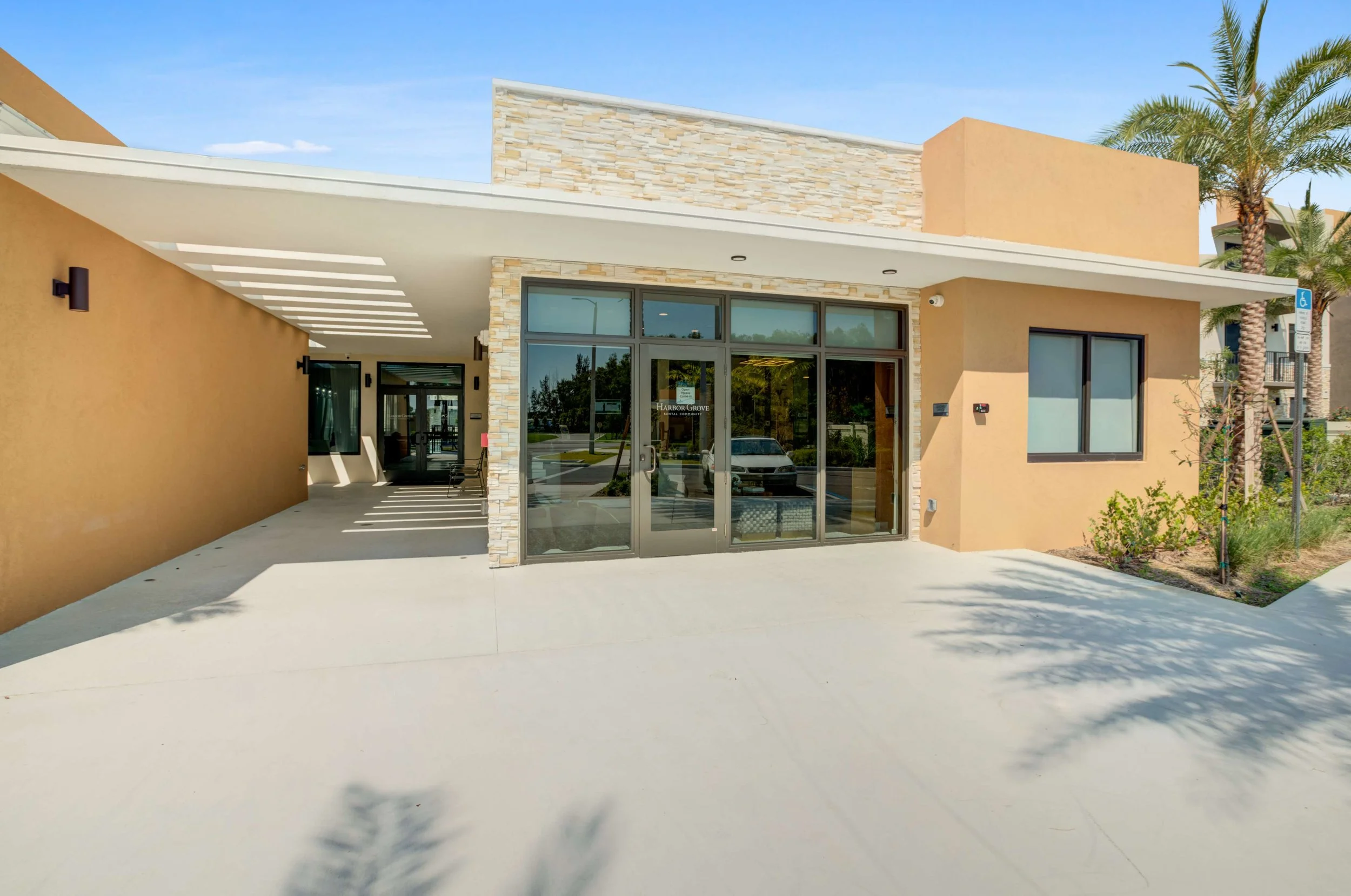 Modern building with beige walls, glass front doors, and a small garden with palm trees outside in sunny weather.