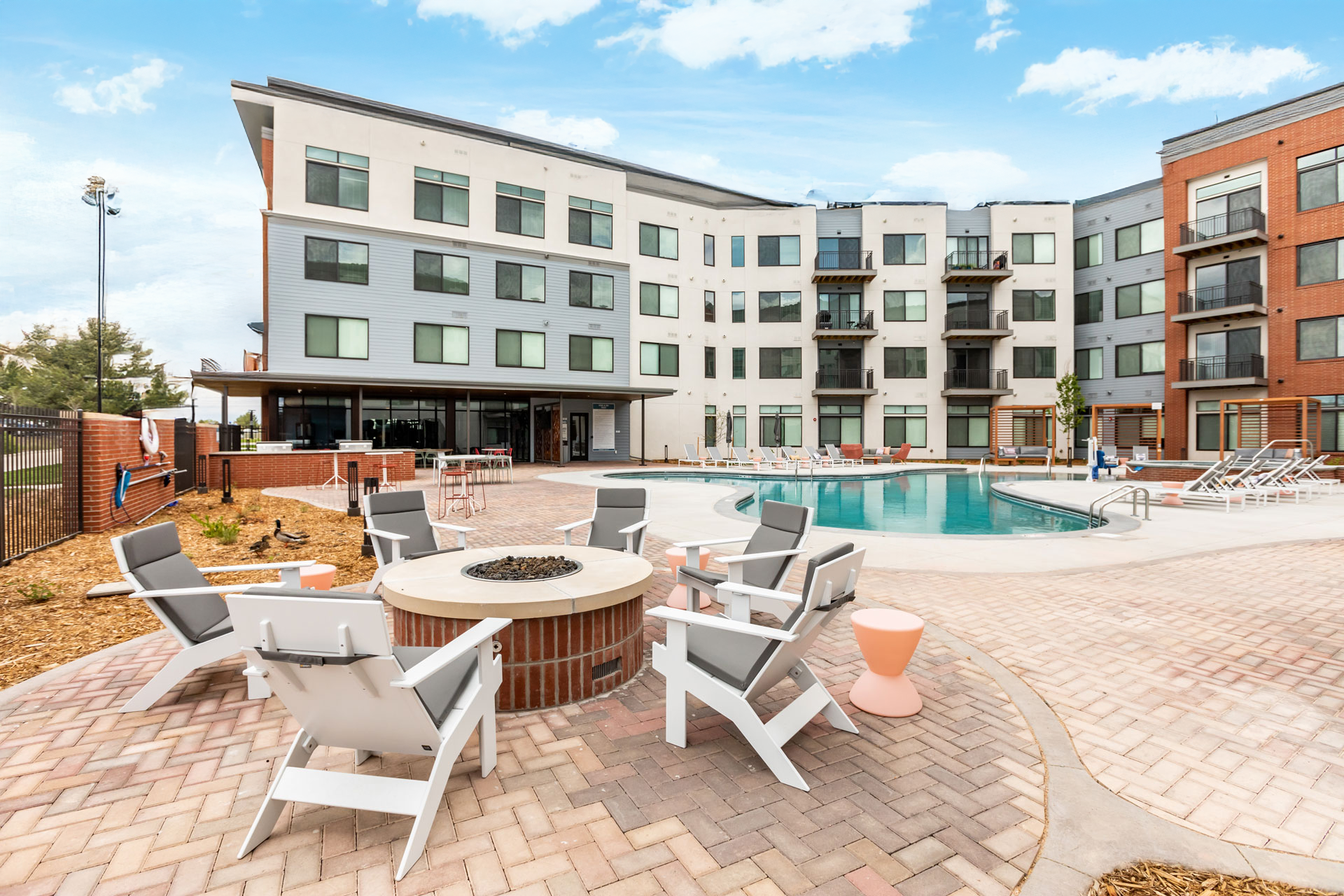 Outdoor swimming pool area in a modern apartment complex with lounge chairs, patio seating, fire pit, and surrounding brick pavement.