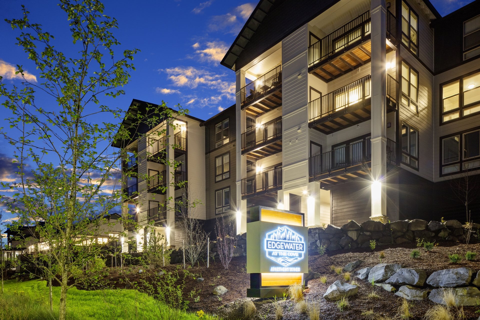 Nighttime exterior view of Edgewater at the Cove Apartments, a multi-story residential building with lit balconies, surrounded by landscaping and a neon sign.