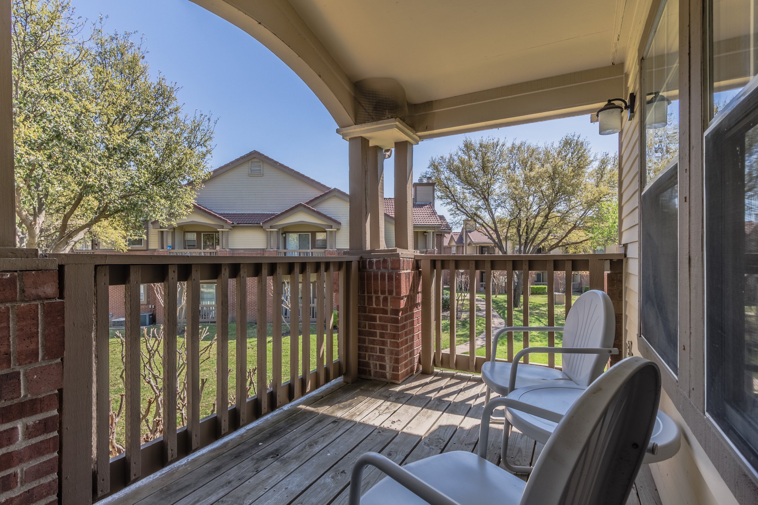 Balcony with two white plastic chairs overlooking a grassy courtyard with trees and neighboring buildings, in sunny weather.