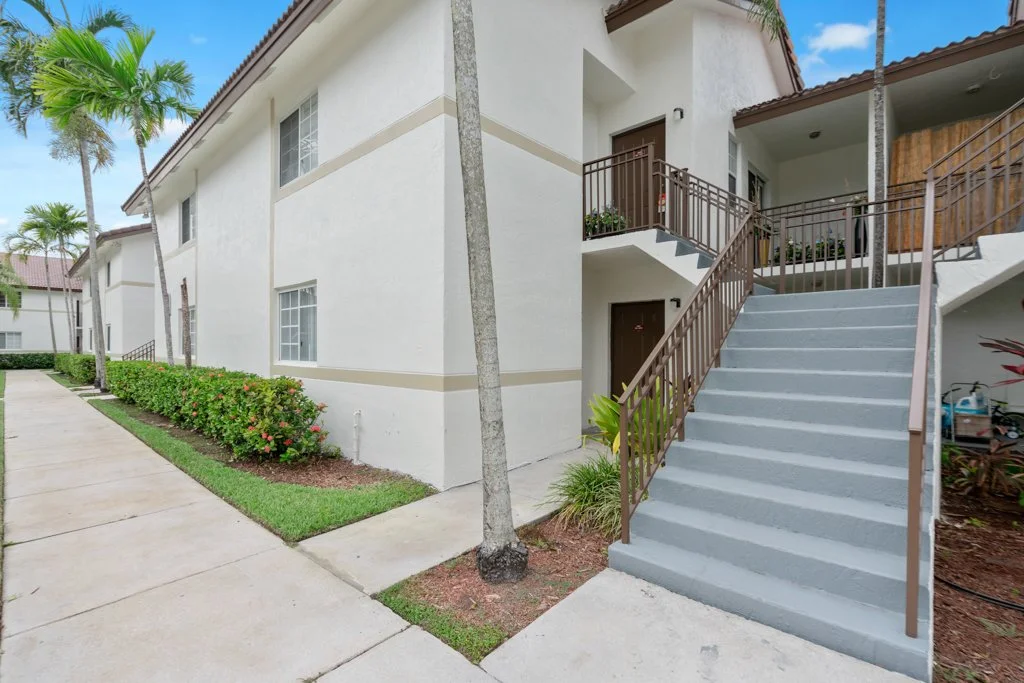 Exterior of a white residential building with stairs, palm trees, and landscaped bushes.