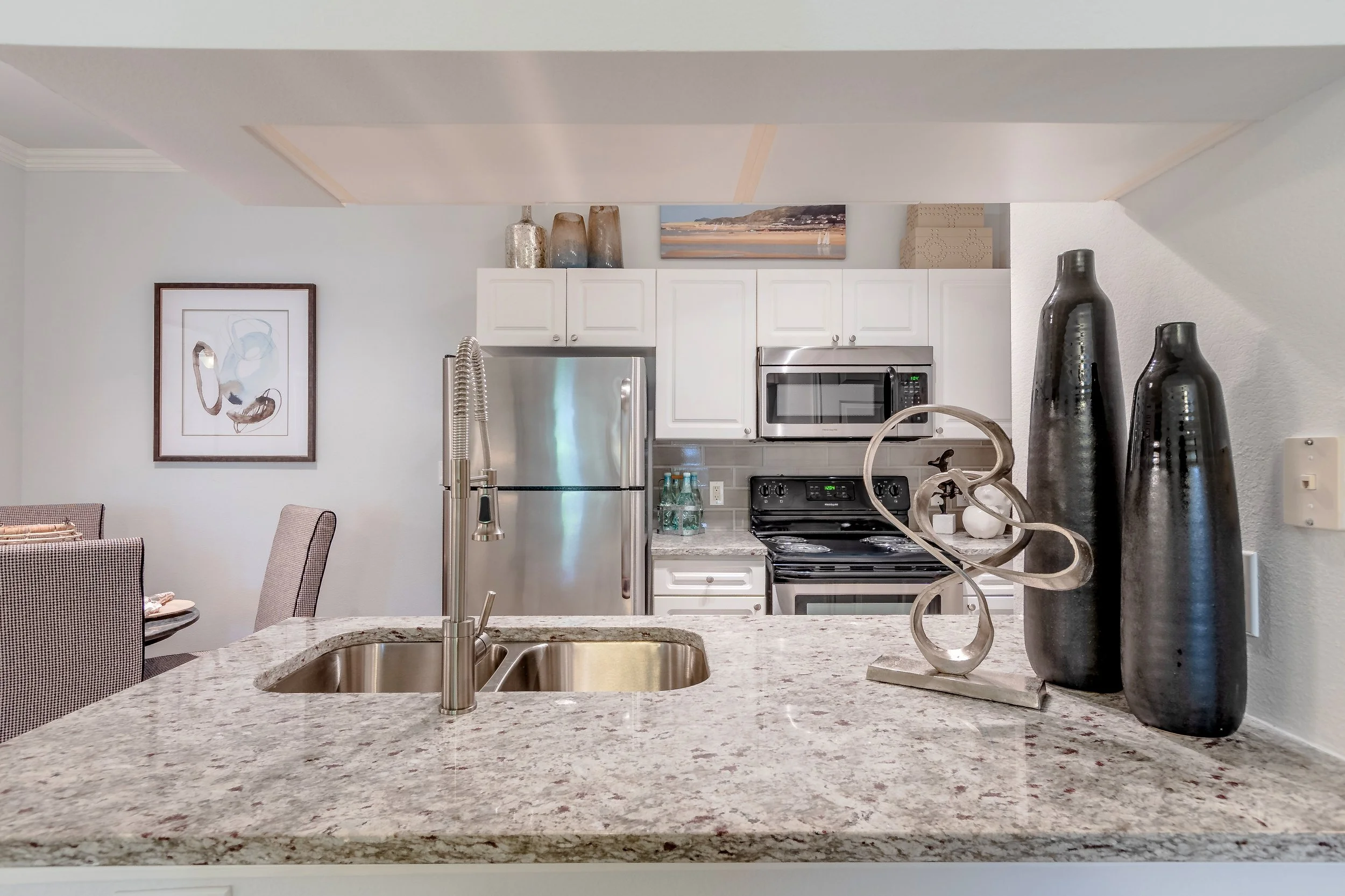 Kitchen with white cabinets, stainless steel refrigerator, oven, microwave, granite countertop island, decorative vases, and artwork on the wall.