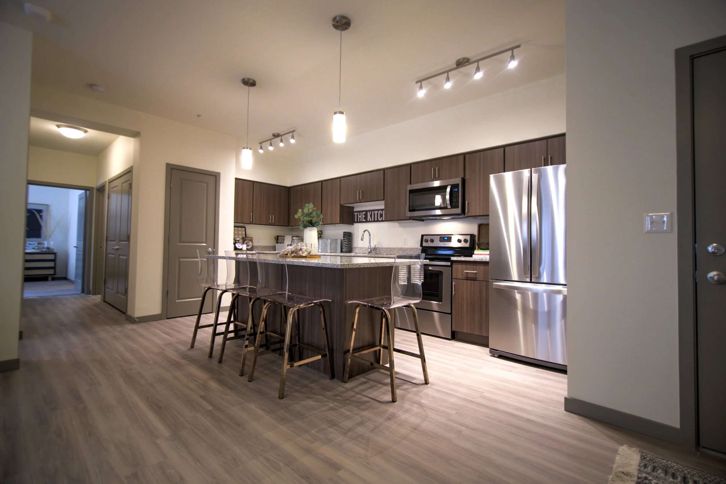 Modern kitchen with dark wood cabinets, stainless steel appliances, a center island with bar stools, pendant and track lighting, a vase with greenery, and a sign reading 'THE KITCHEN'.