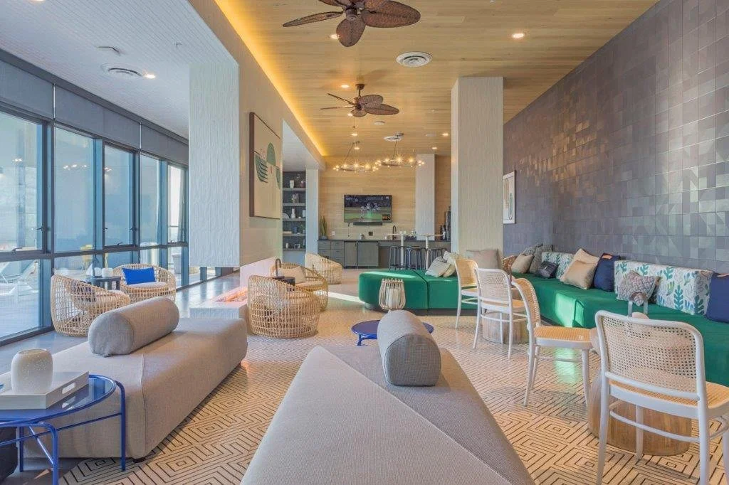 Modern living room with large windows, wicker chairs, a green sofa with pillows, and a kitchen area in the background.
