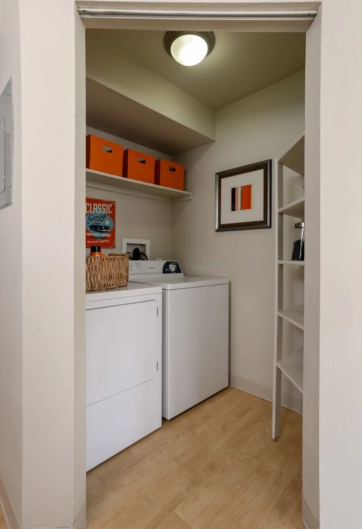A laundry room with a washing machine and dryer, a basket on top, orange storage boxes on a shelf, a framed art piece on the wall, and a built-in shelving unit to the right.