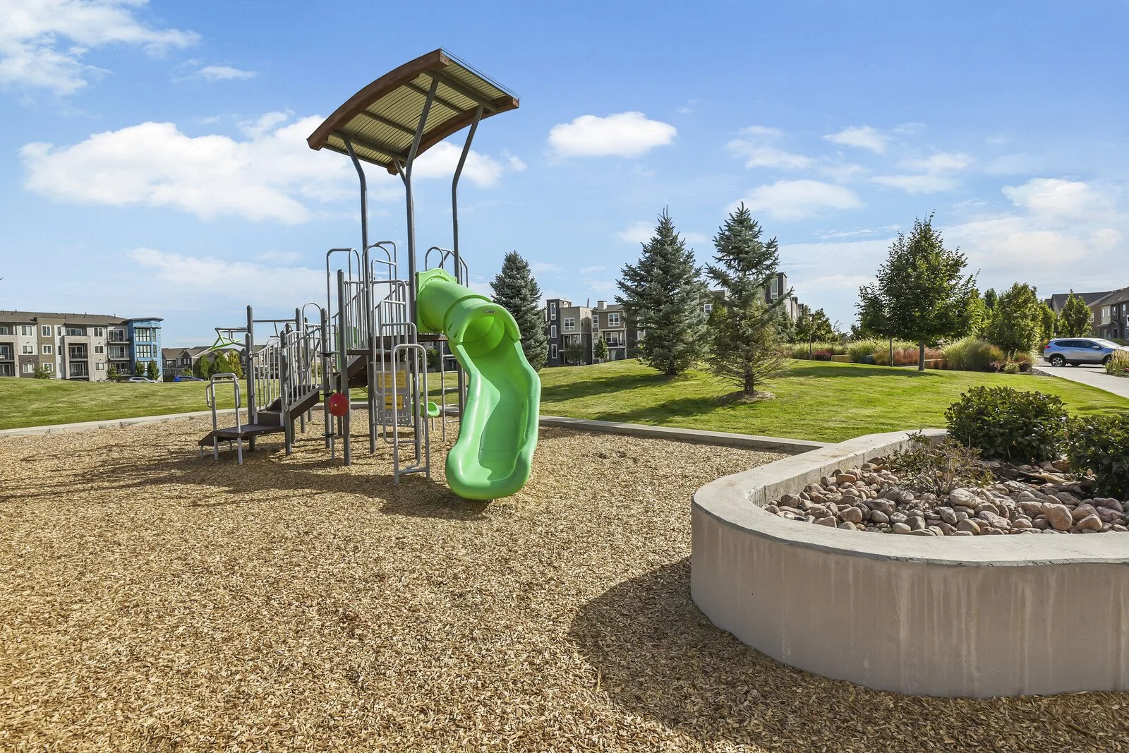 Childrens playground with green slide and climbing structure in a park setting with trees and apartment buildings in the background.