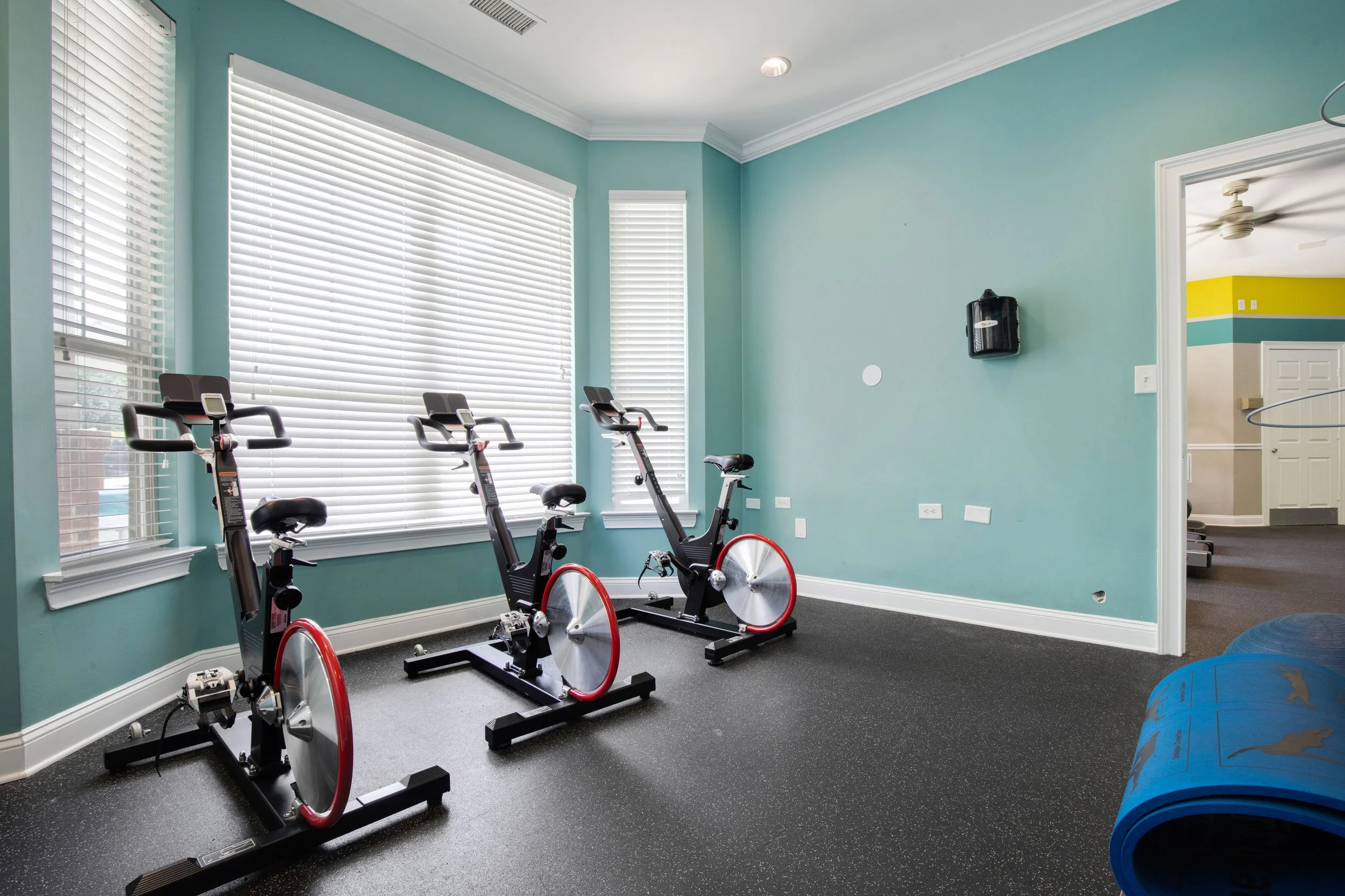 Room with two stationary exercise bikes and a blue exercise mat on black flooring, with windows covered by white blinds and a doorway leading to another room with yellow and teal walls.