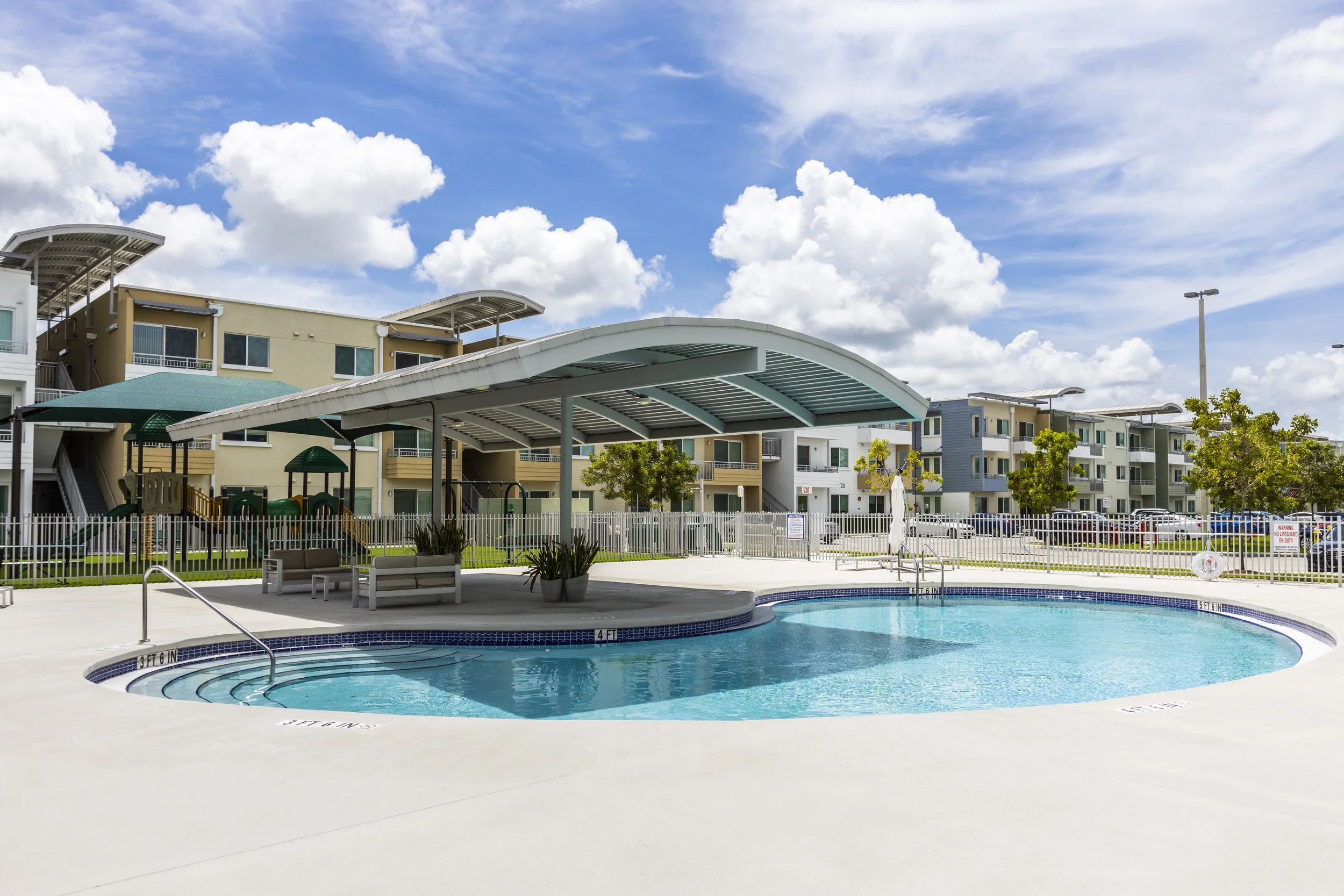 Community swimming pool with a covered seating area, playground, and apartment buildings in the background under a partly cloudy sky.