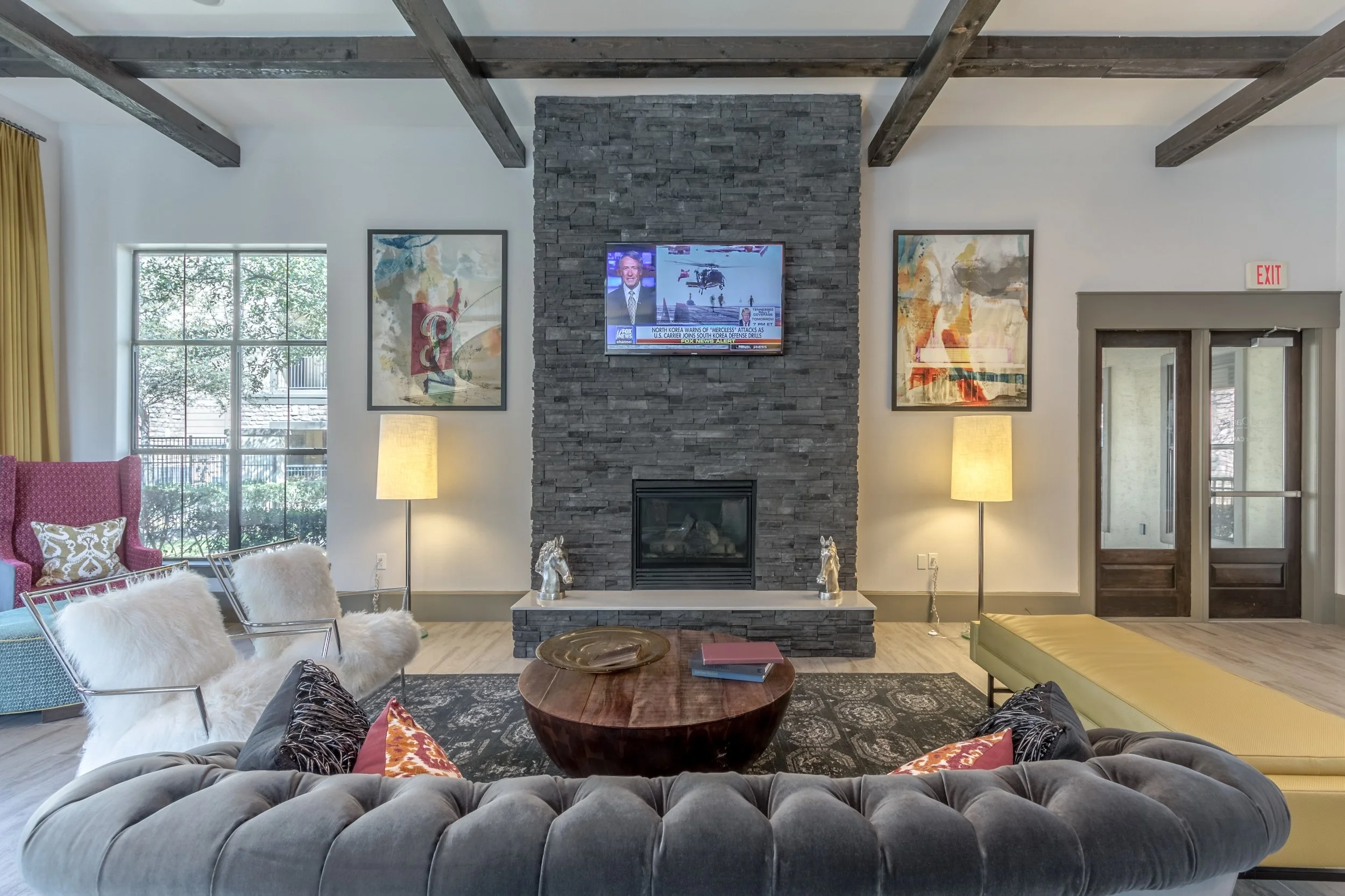 Living room with a fireplace on a stone wall, two framed artworks, a mounted television, two lamps, a large window with outside greenery, a pink armchair with a patterned pillow, a teal armchair, two white furry chairs, a round wooden coffee table wi