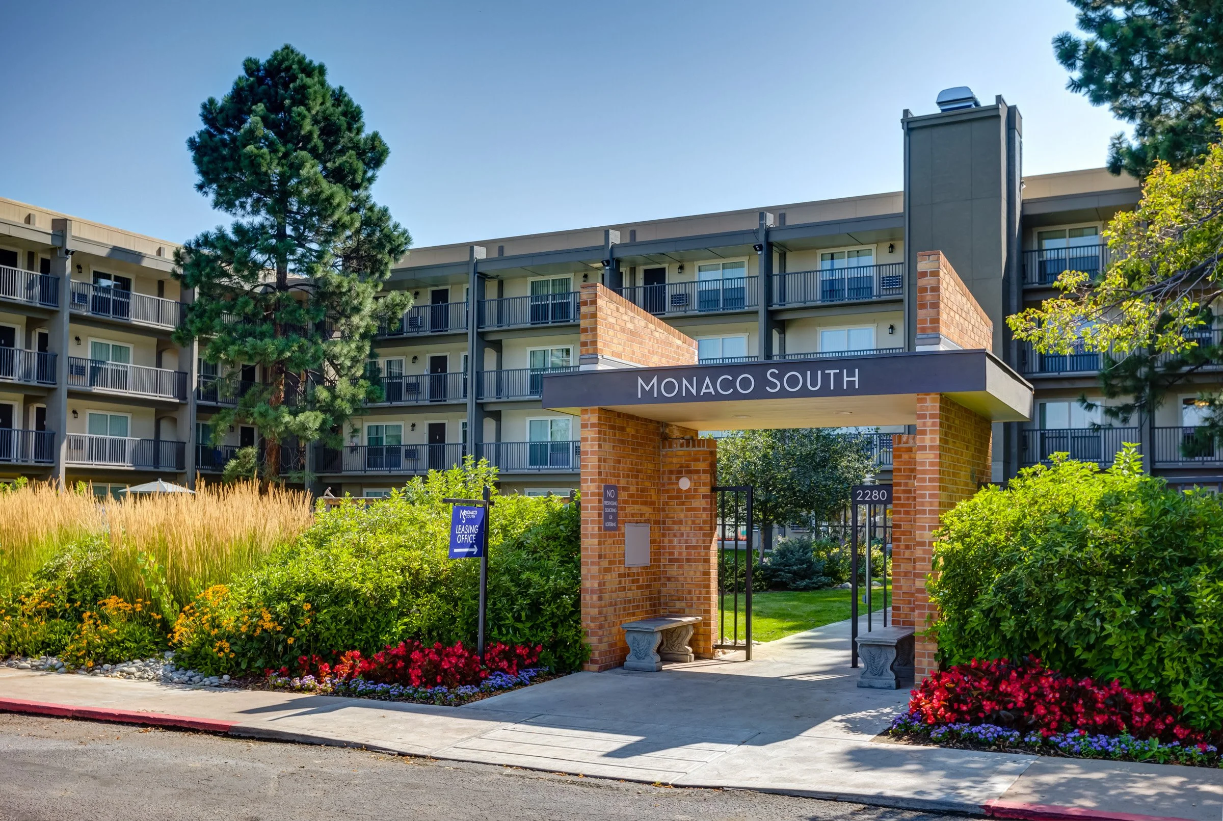 Apartment complex entrance with a sign reading "Monaco South", surrounded by landscaped bushes and flowers, and a building with multiple balconies in the background.