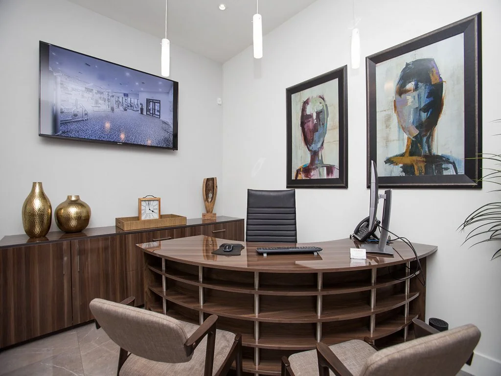 Office with a wooden desk, black leather chair, two beige chairs, wall art portraits, a wall-mounted TV, and decorative vases.