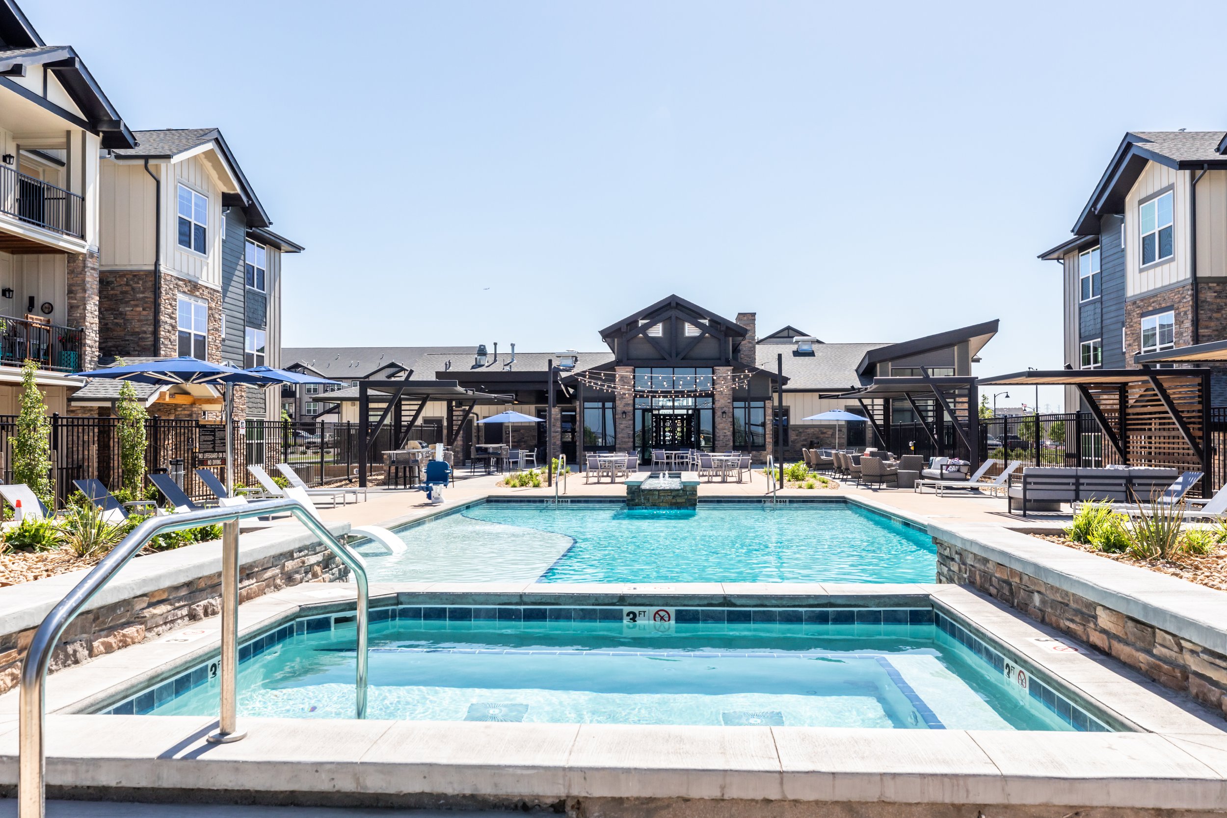 Apartment complex outdoor swimming pool area with hot tub, lounge chairs, umbrellas, and a clubhouse in the background under a clear blue sky.