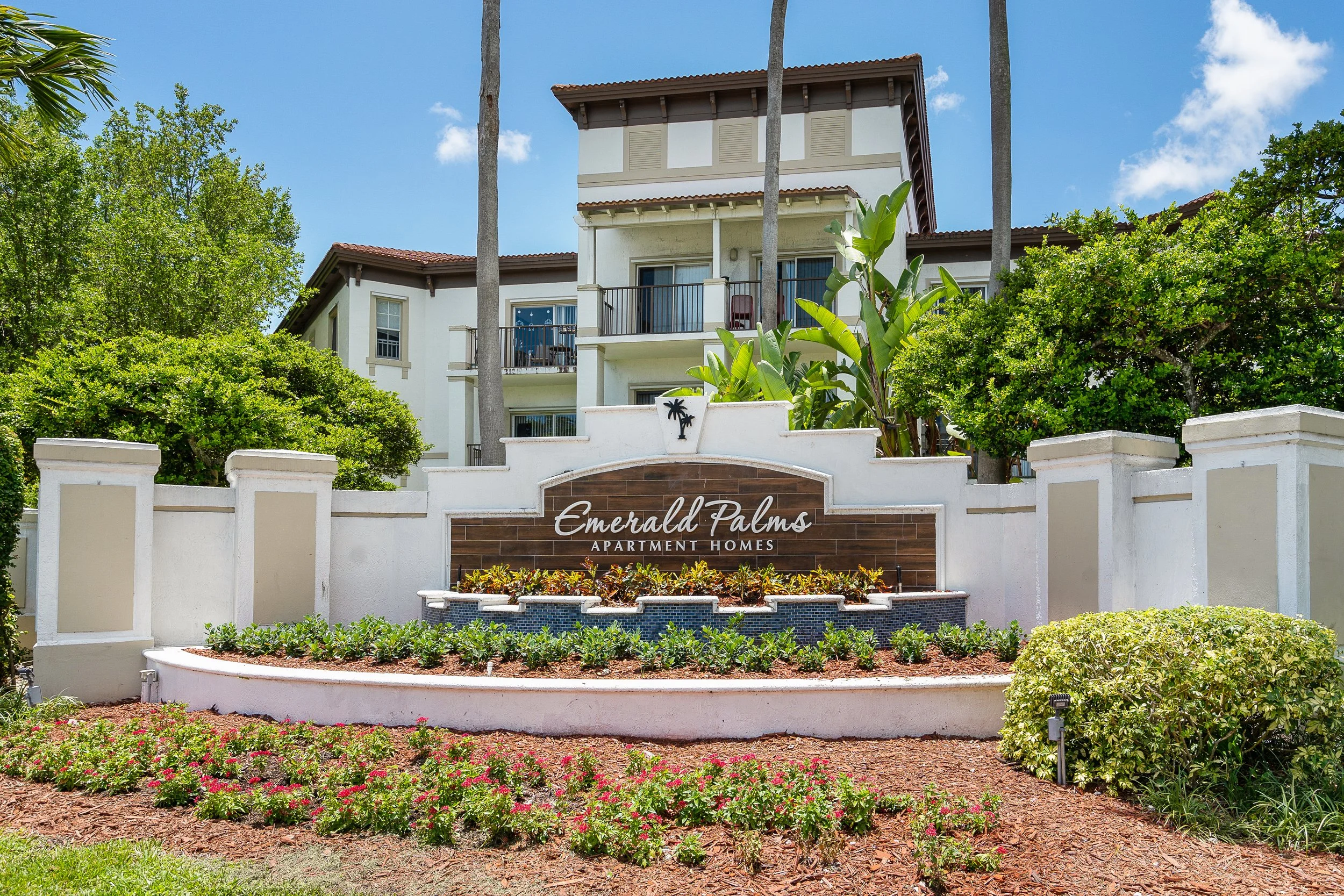 Entrance sign for Emerald Palms apartment homes with landscaping and greenery in front of a multi-story residential building, under a blue sky.