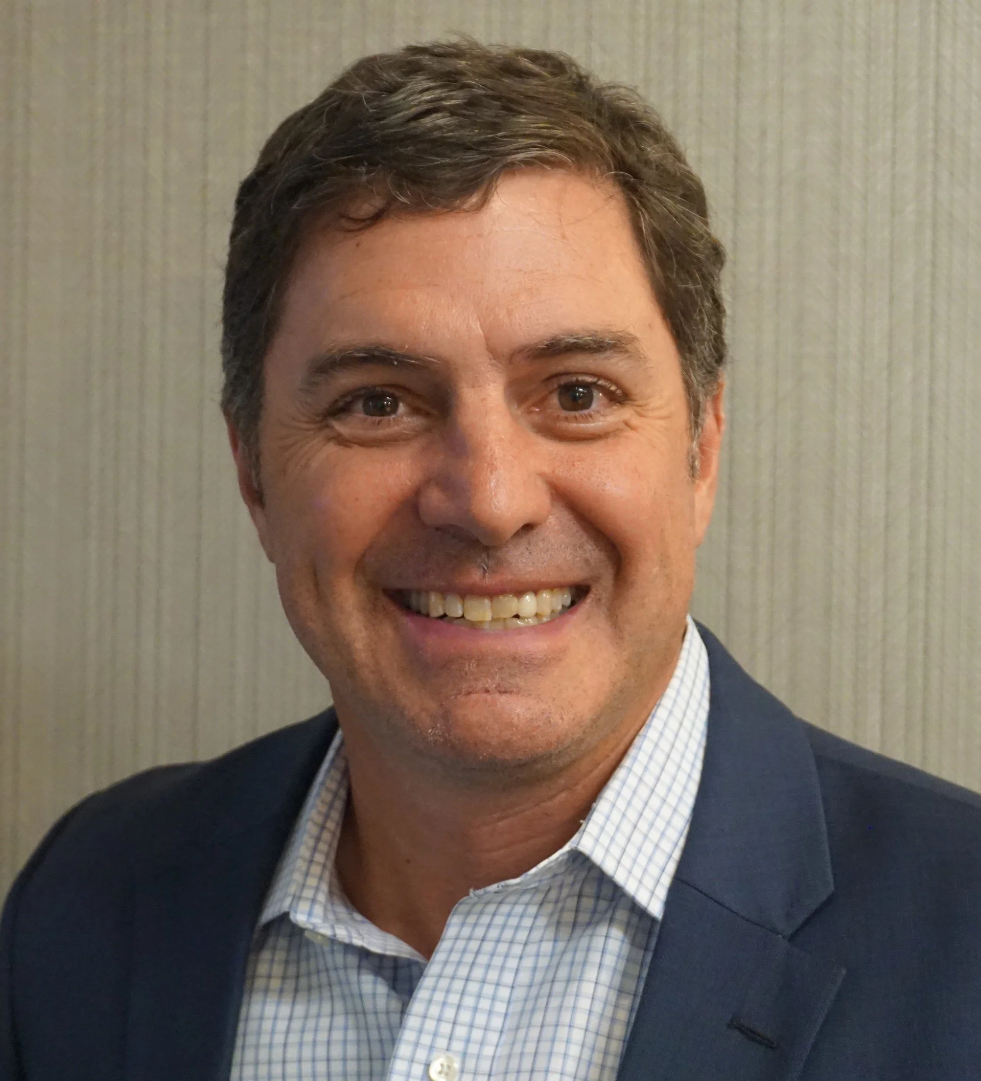 A smiling man with dark curly hair, wearing a white shirt and blue blazer.