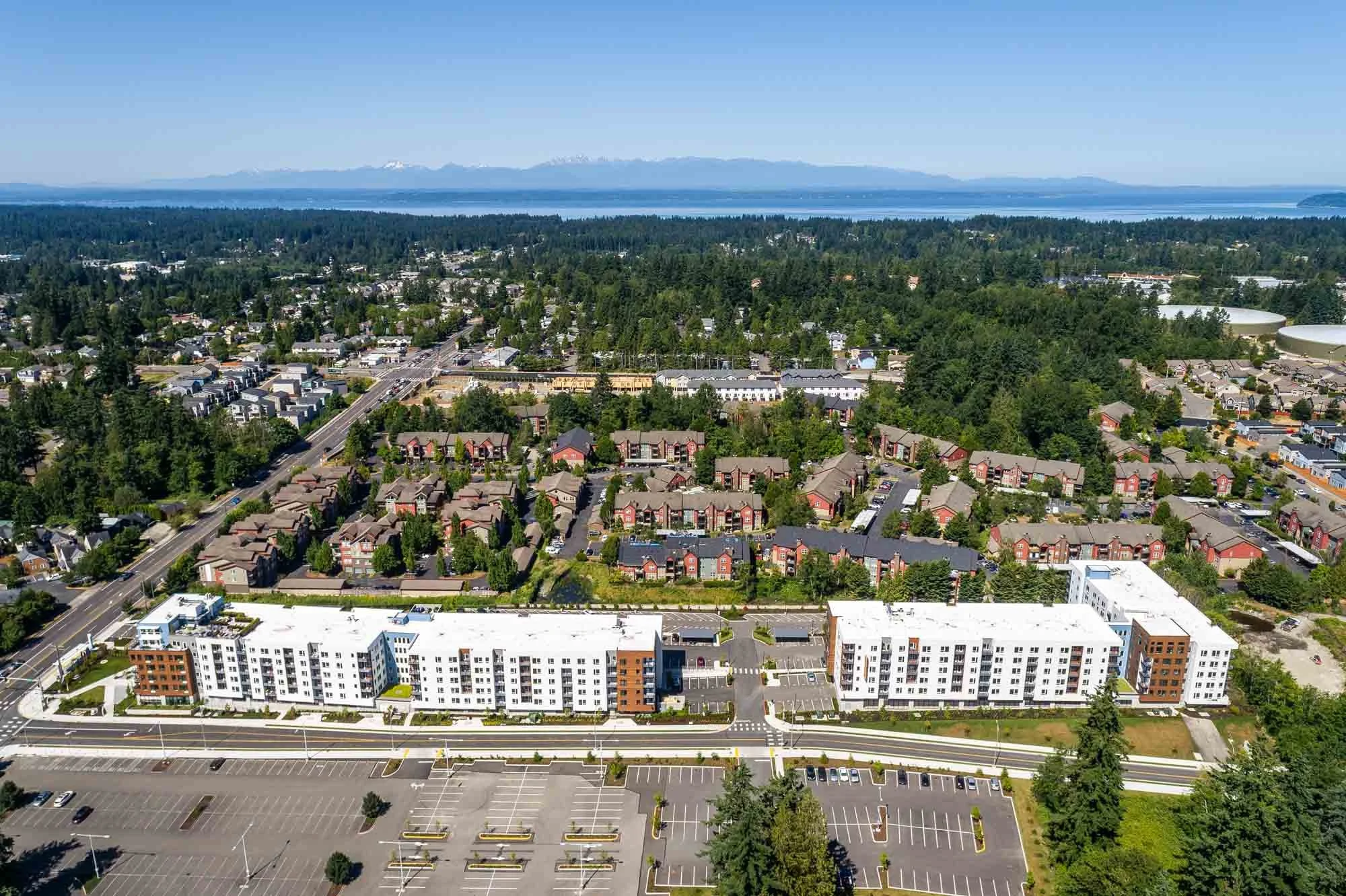 Aerial view of a cityscape with a large white apartment building in the foreground, surrounded by parking lots and streets. In the background, there are residential neighborhoods with trees, hills, and a body of water with distant mountains.