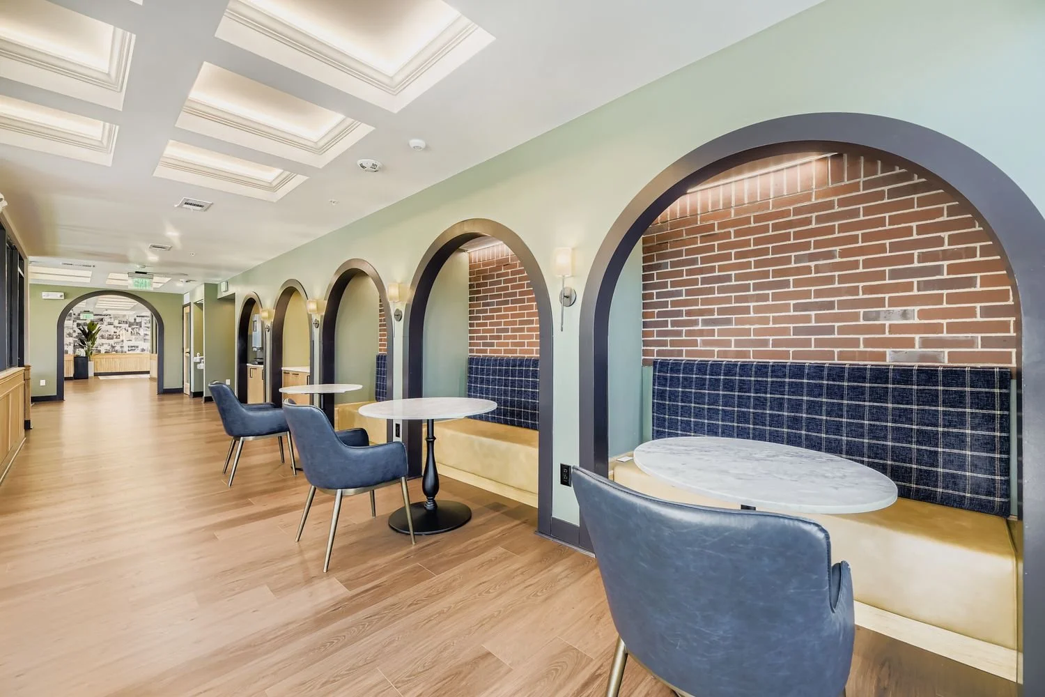 Interior of a modern cafe with round tables, blue chairs, brick wall niches with plaid fabric panels, green walls, and wooden flooring.