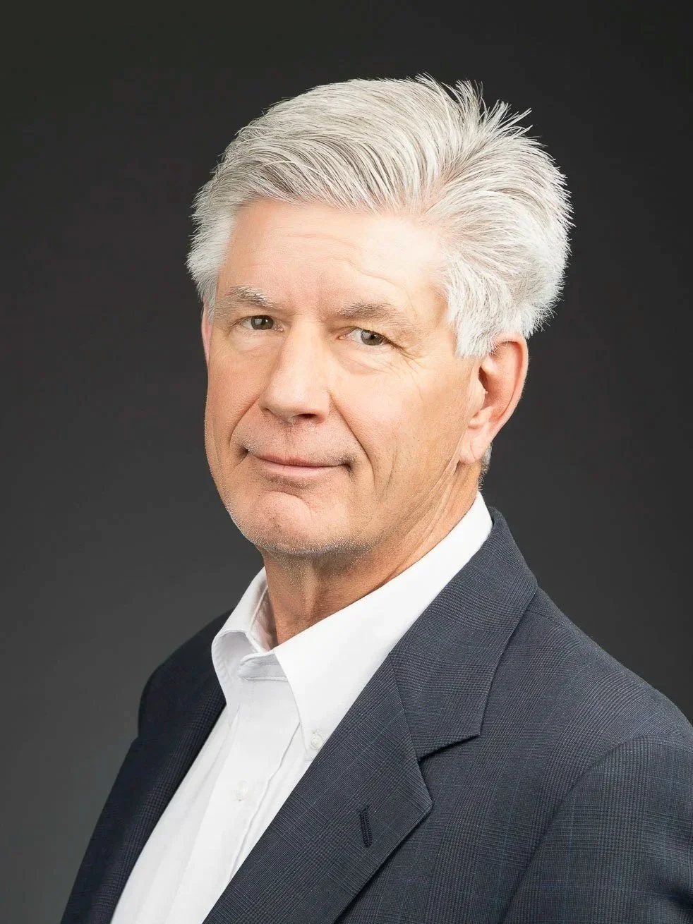 A portrait of a mature man with gray hair wearing a dark suit and white shirt, standing against a dark background.