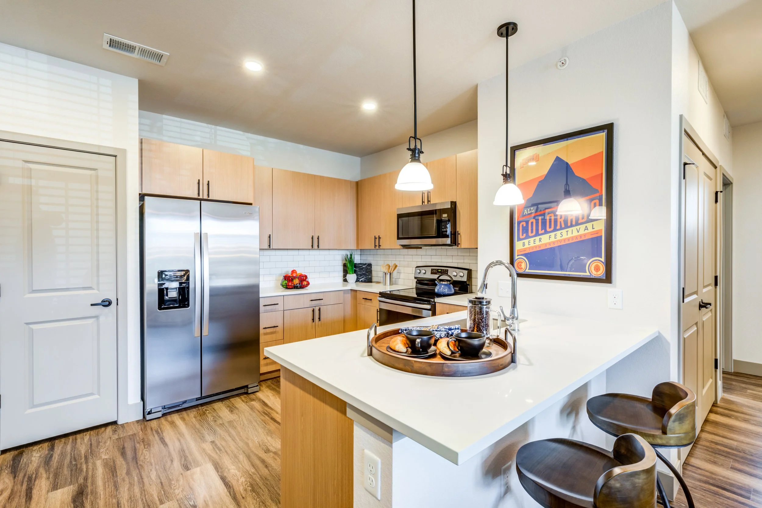 Modern kitchen with light wood cabinets, stainless steel refrigerator, oven, microwave, and white subway tile backsplash. There is a white countertop with a tray of breakfast items and bar stools at the counter. A colorful poster is on the wall.