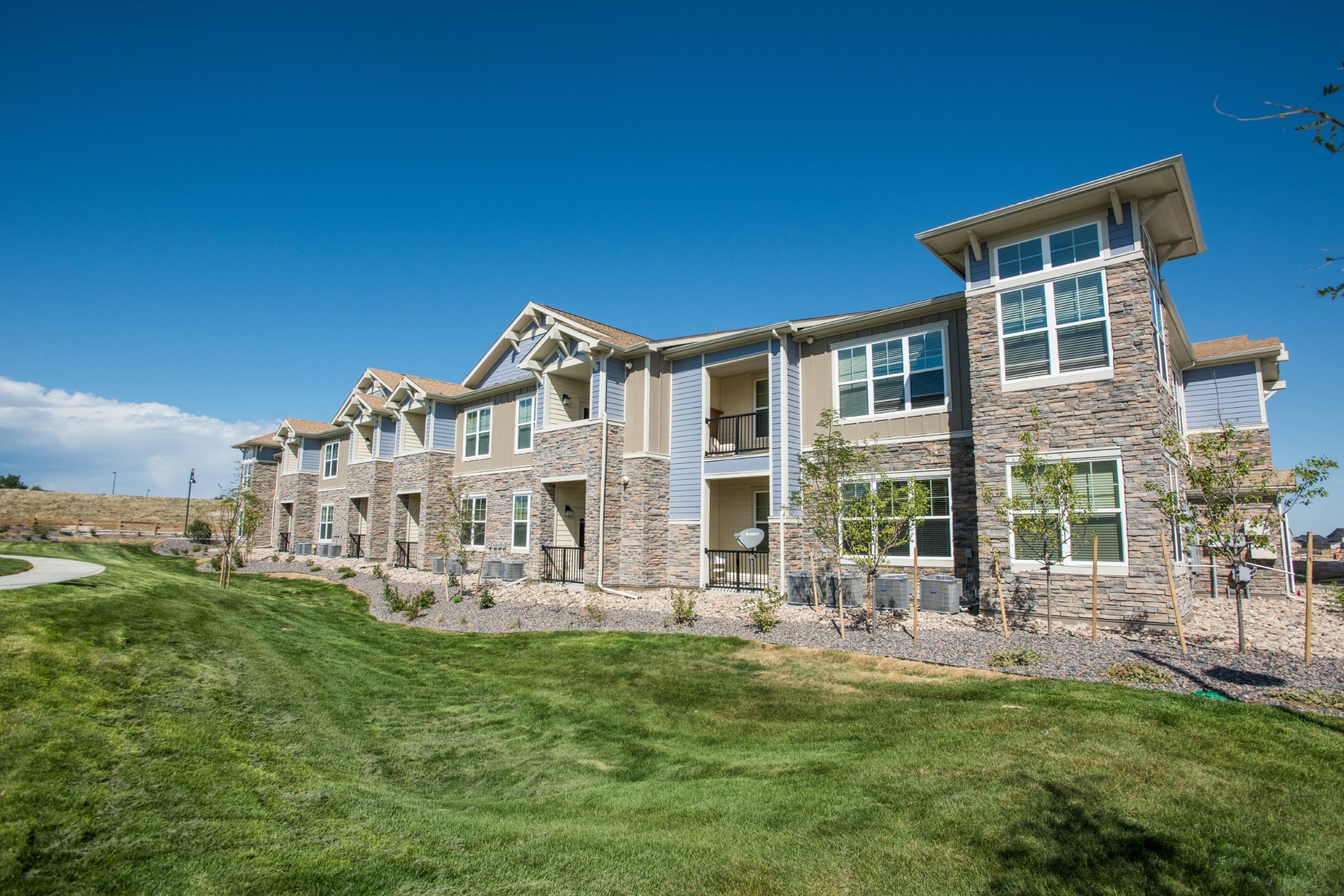 Multi-story residential buildings with stone and siding exteriors, surrounded by green lawn and small trees, under a clear blue sky.