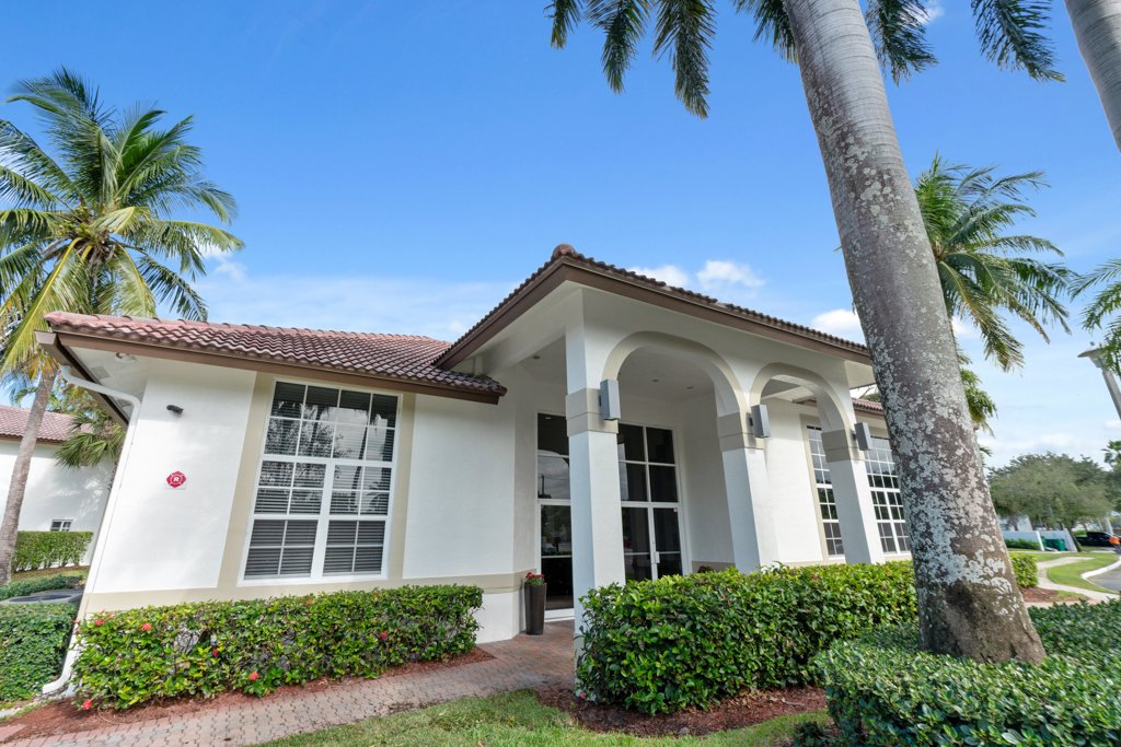 A white house with large windows, a tiled roof, and a covered entrance, surrounded by palm trees and trimmed bushes, under a clear blue sky.