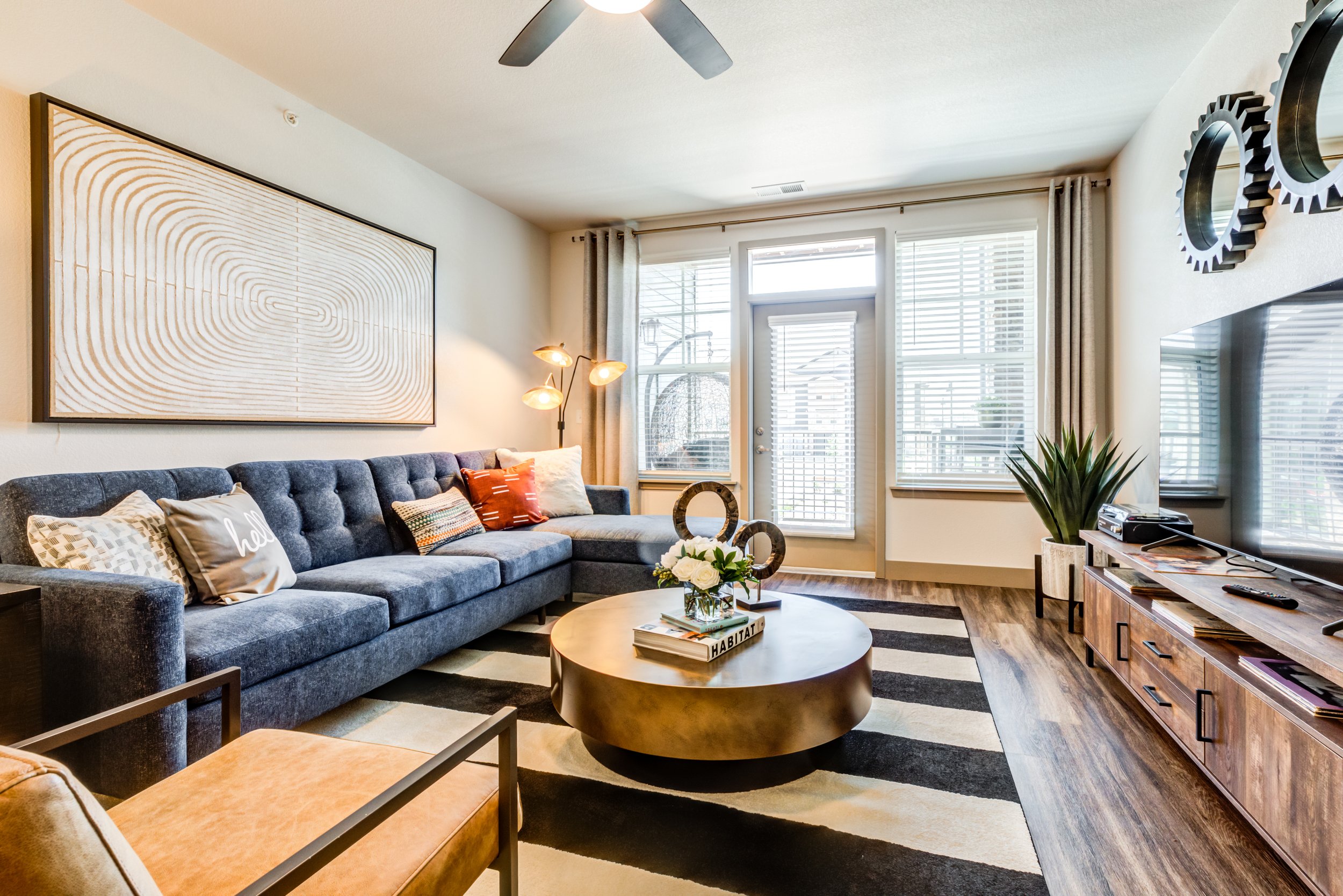 Living room with a dark gray sofa, a round gold coffee table, a striped black and white rug, and wooden flooring. There are various pillows on the sofa, a large wall art piece above, and a flat-screen TV with a wooden console. A window, a glass door 