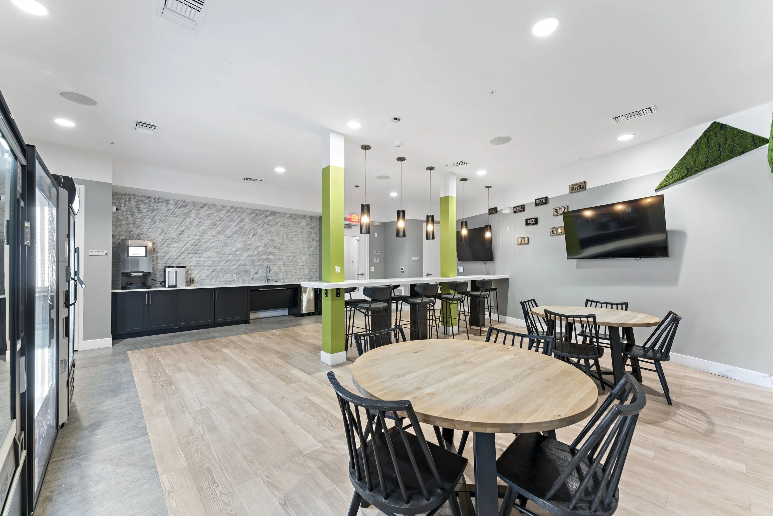 Modern lounge area with tables and chairs, a bar with high stools, a coffee station, and a TV on the wall.