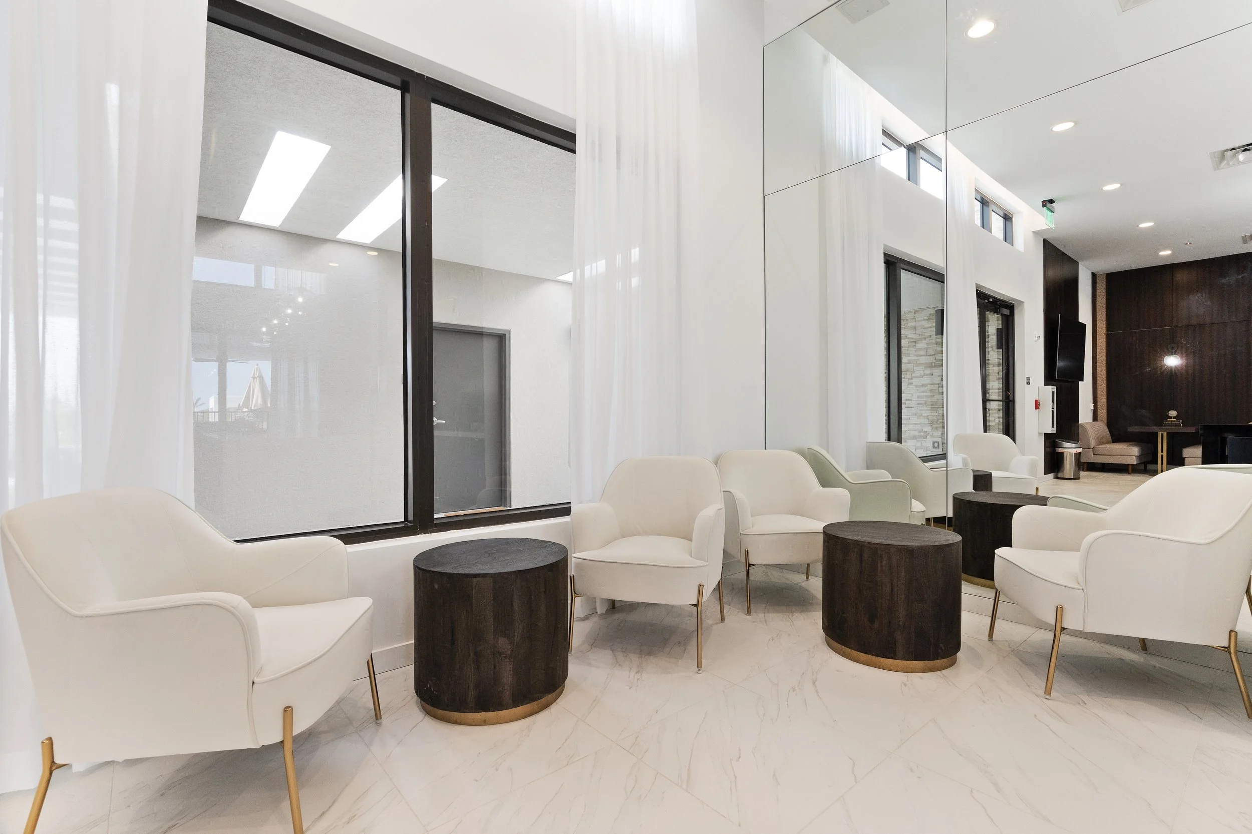 Modern waiting area with white armchairs, dark circular coffee tables, large windows with white curtains, and minimalist decor.