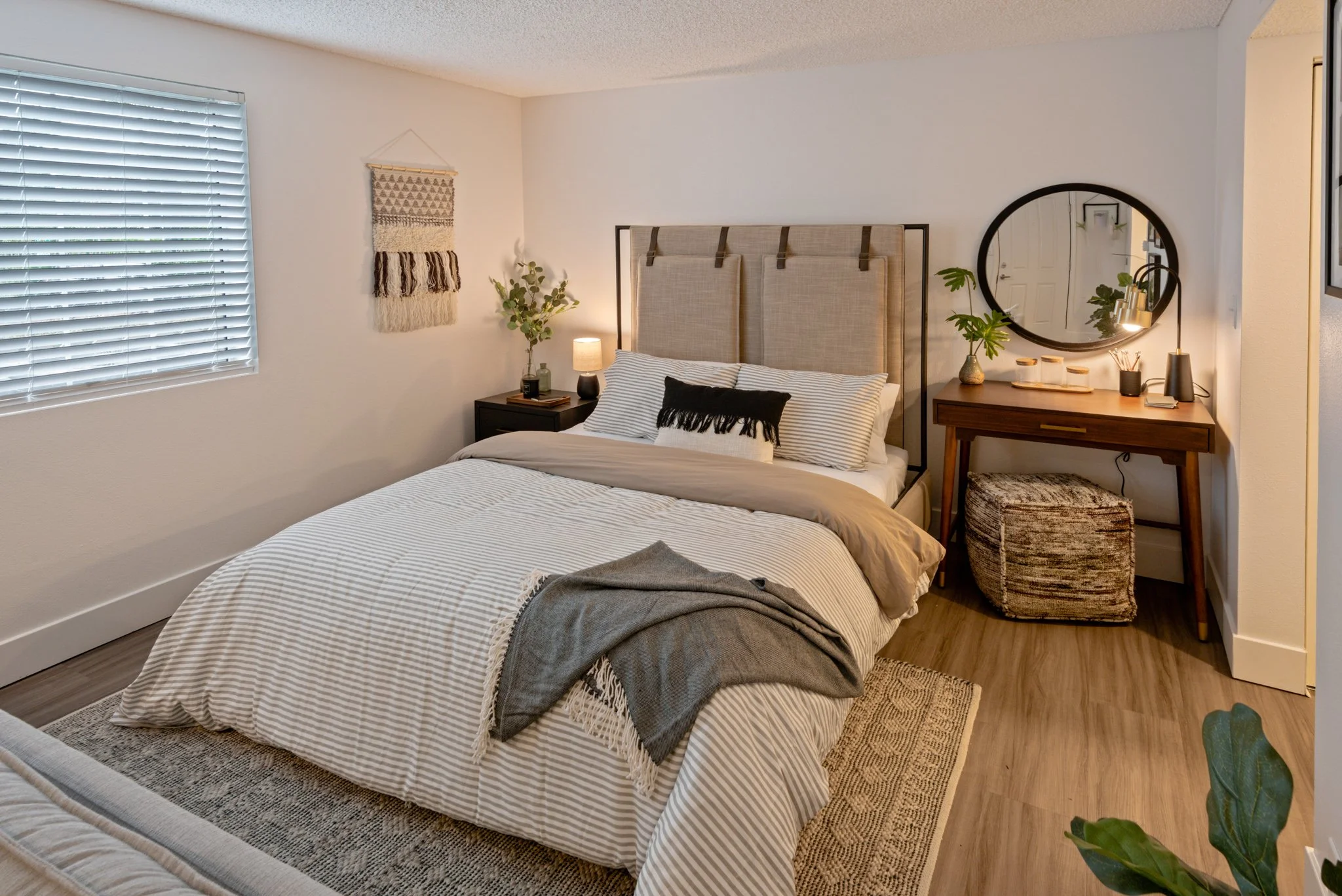 A neatly made bedroom with a bed featuring striped bedding and a black throw pillow. The room has a black bedside table with a small lamp and decorative vase with greenery. There’s a wooden desk with a round mirror, various decor items, and a small w