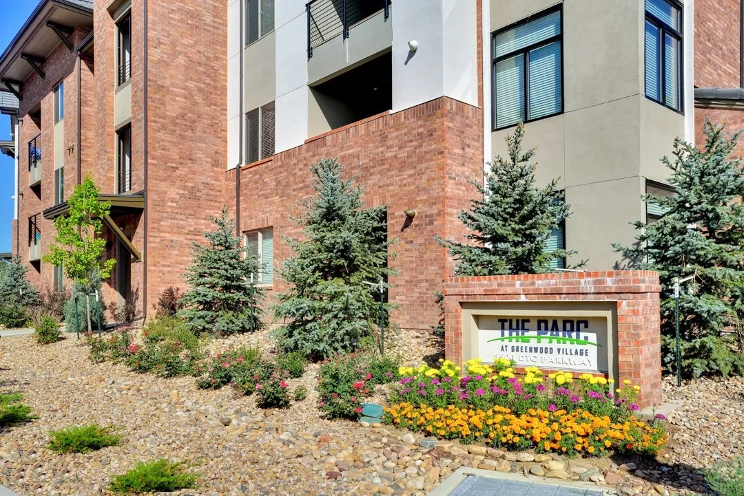 Residential building with brick and beige exterior, surrounded by landscaped garden with flowers and small pine trees, and a sign that reads 'THE PARC at Greenwood Village'.