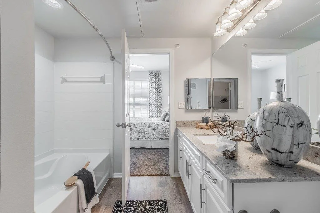 Bathroom with white vanity and granite countertop, large decorative vases, mirror, and lights above. View into a bedroom through the open door, with a bed and window.