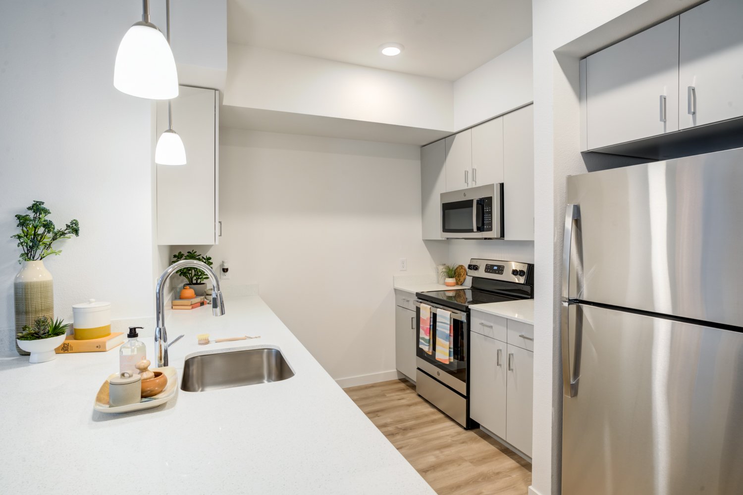 Modern kitchen with white countertops, stainless steel refrigerator, microwave, and oven, decorated with potted plants, towels, and wooden accents.
