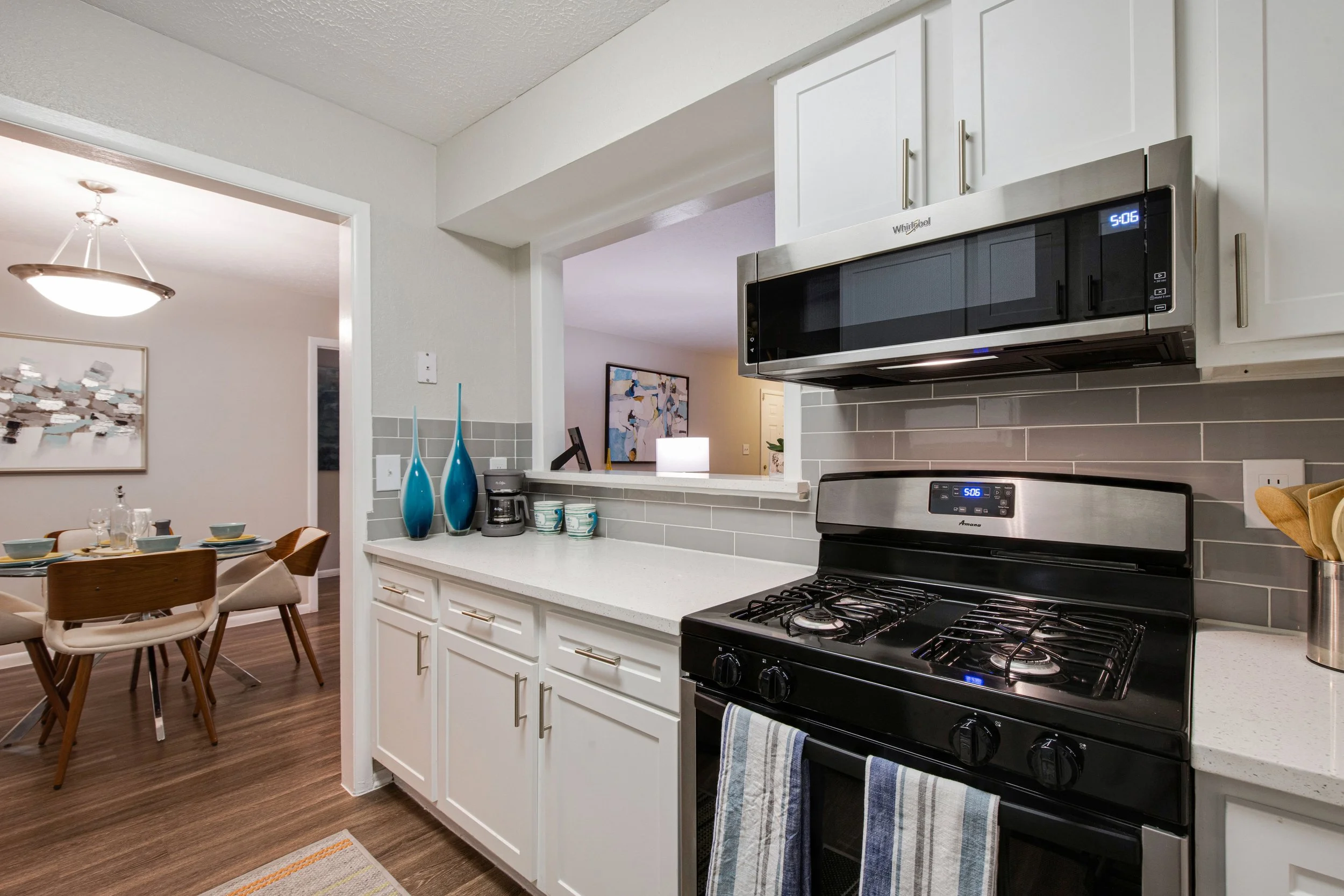 Modern kitchen with white cabinets, gray backsplash, black stove, and microwave. The kitchen opens into a dining area with a wooden table and six chairs, decorated with a large abstract painting and a hanging light fixture.