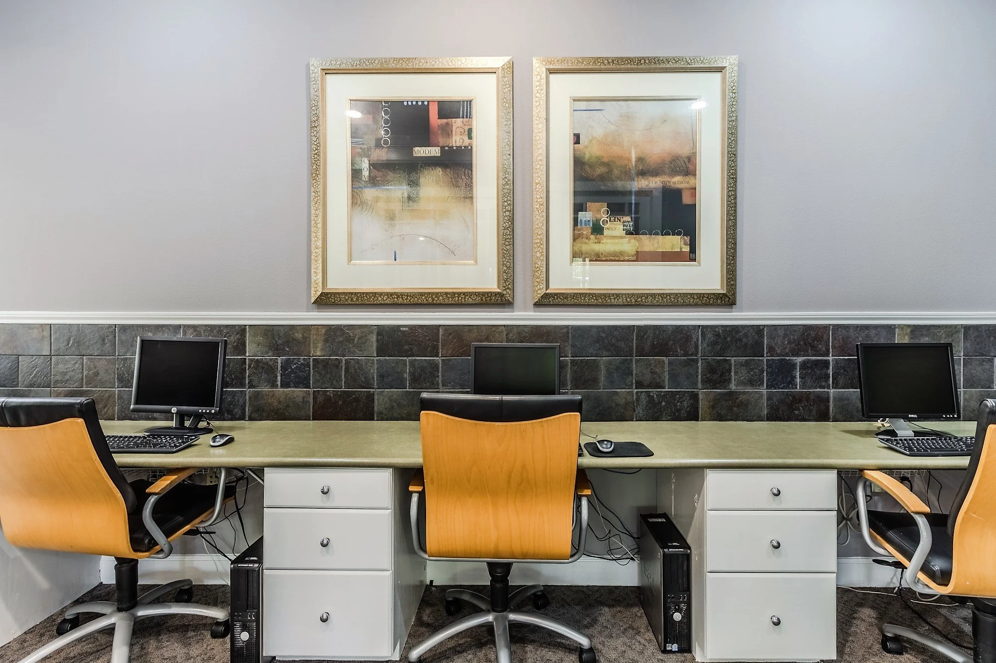 An office workspace with three computer desks, each equipped with a monitor, mouse, and keyboard. Two desks have black chairs with wood armrests, and the middle desk has a tan-colored chair. There are wall art frames hanging above the desks, and the 