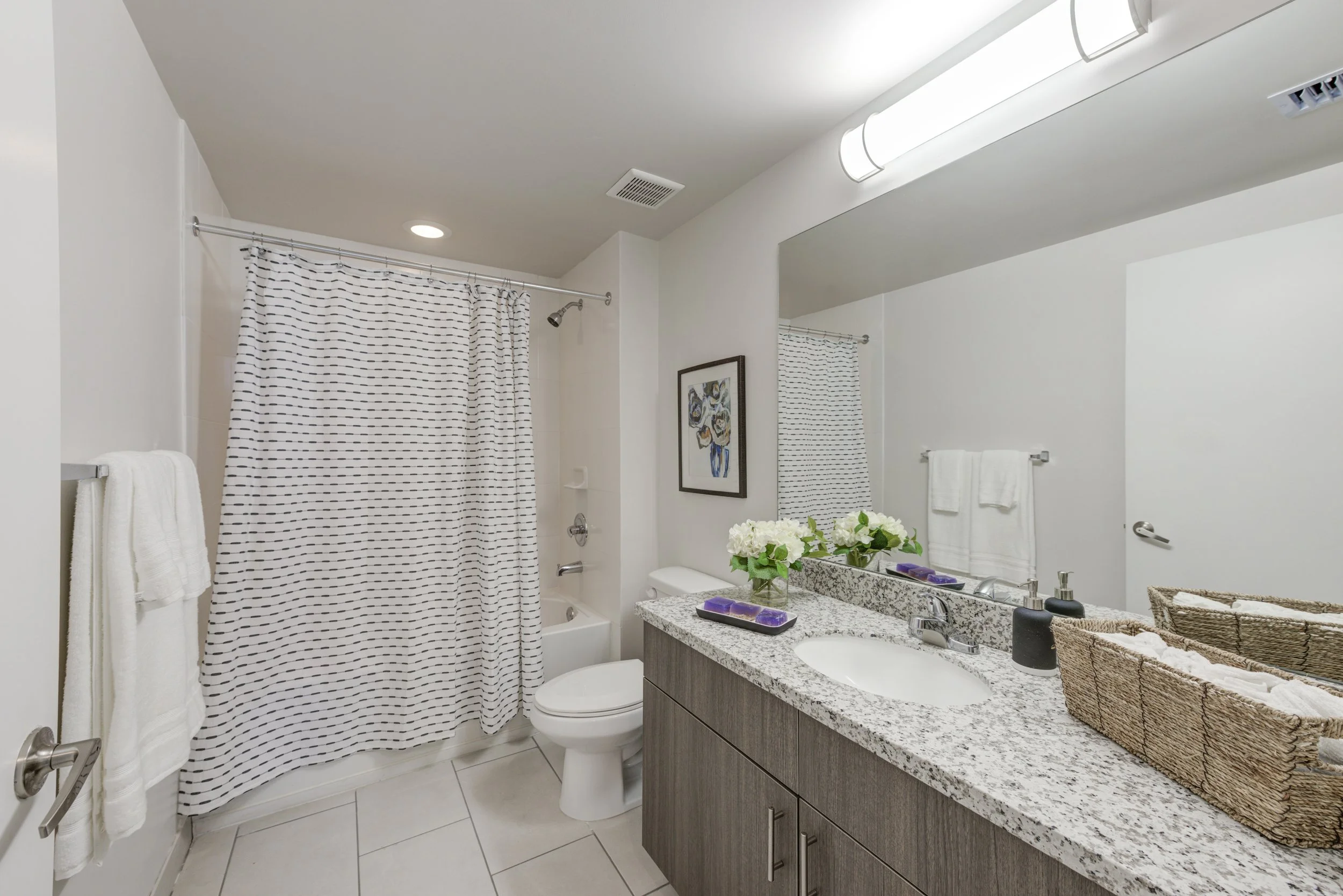 Modern bathroom with a granite countertop vanity, large mirror, shower with striped curtain, toilet, framed artwork, and flower arrangement.