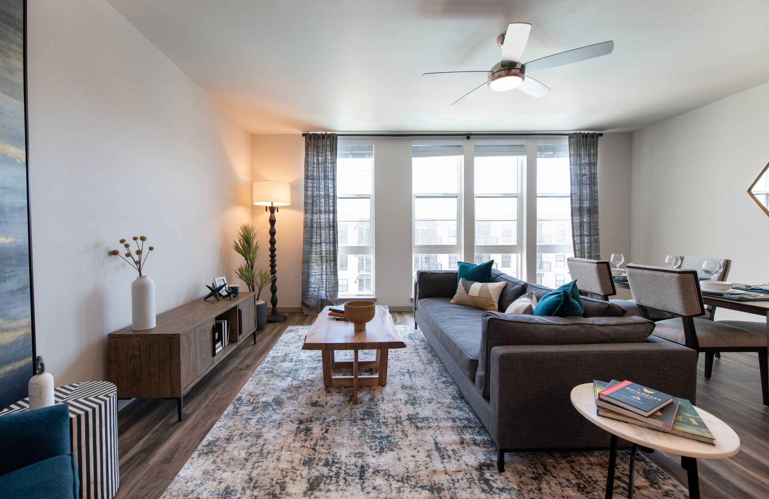 Living room with large windows, gray sofa, wooden coffee table, dining table with chairs, rug, potted plants, standing lamp, ceiling fan, and decorative wall mirror.