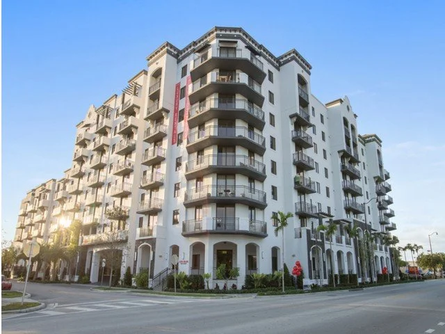 A modern multi-story apartment building with balconies and palm trees along a city street.