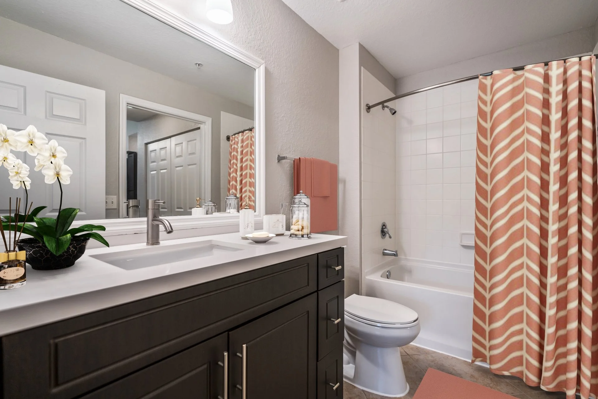 A modern bathroom with a white vanity and dark cabinets, a large mirror, a white toilet, a bathtub with a herringbone patterned shower curtain in pink and white, pink towels, decorative jars on the counter, a potted orchid, and a tiled wall behind th