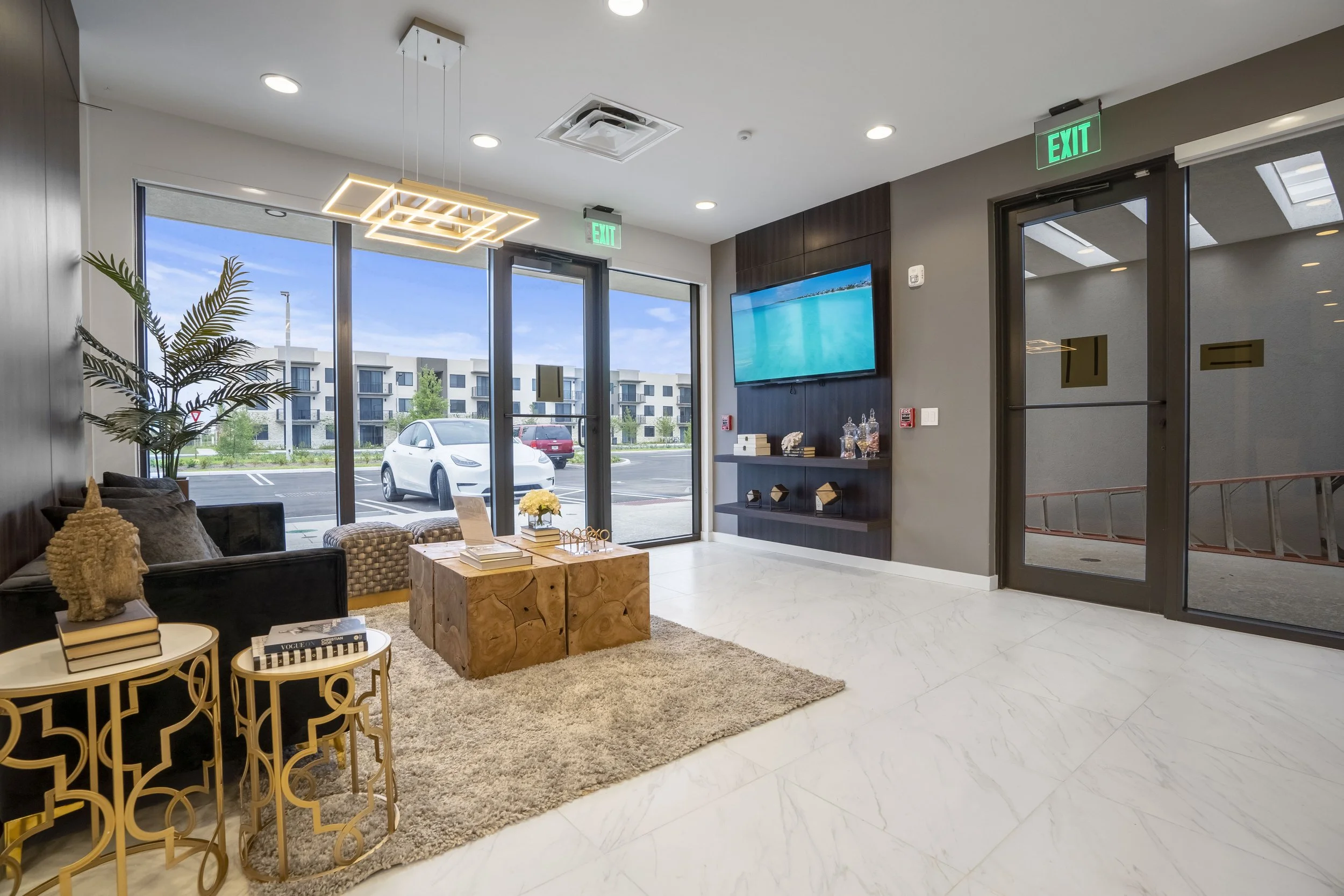 Modern lobby with a seating area, a large window showing parked cars outside, a wall-mounted television, and decorative shelves. The room has polished white floors, contemporary furniture, and ceiling lighting.
