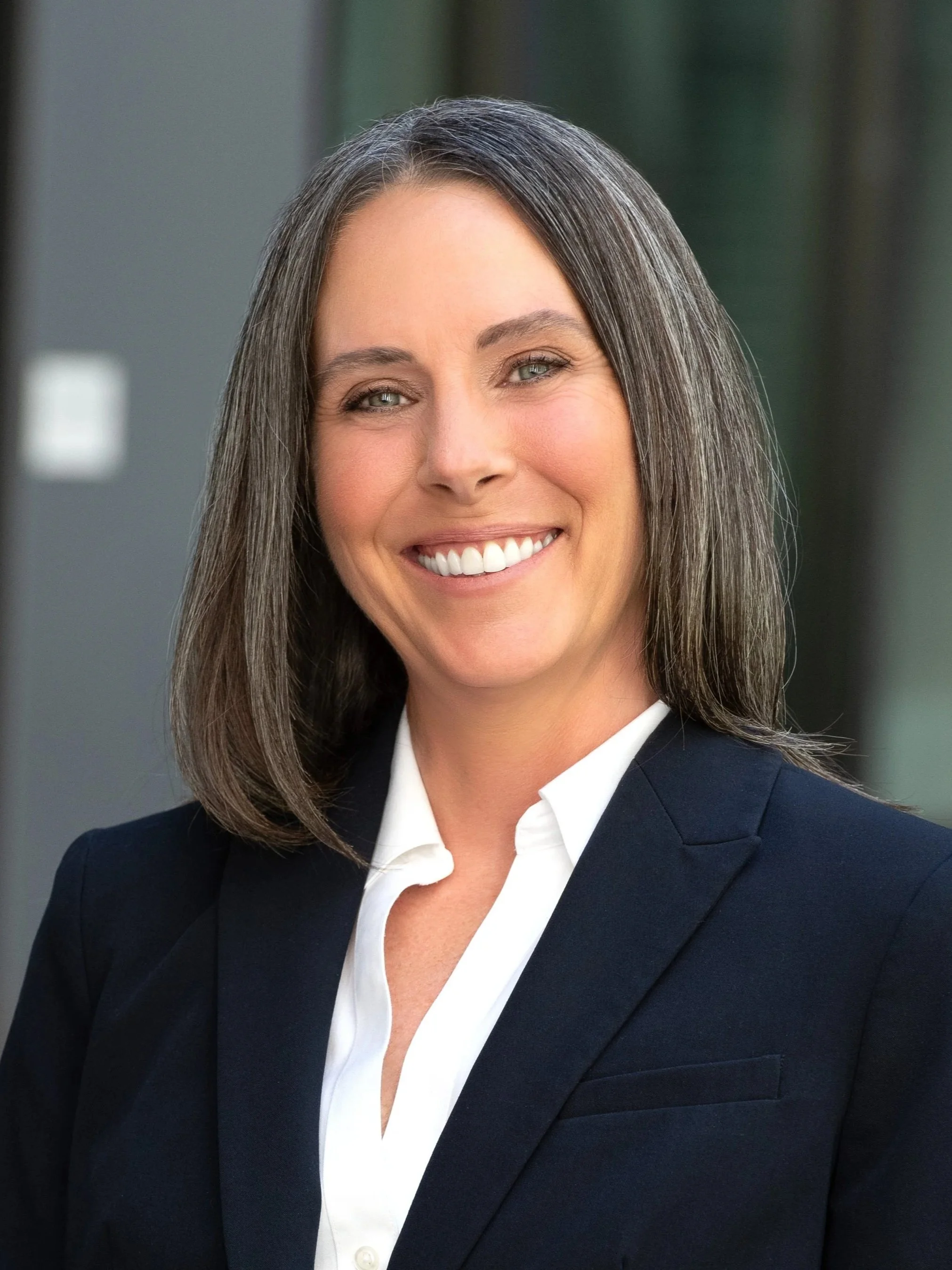 A smiling woman with shoulder-length brown hair, wearing a dark blazer and white shirt, standing in front of an office building.