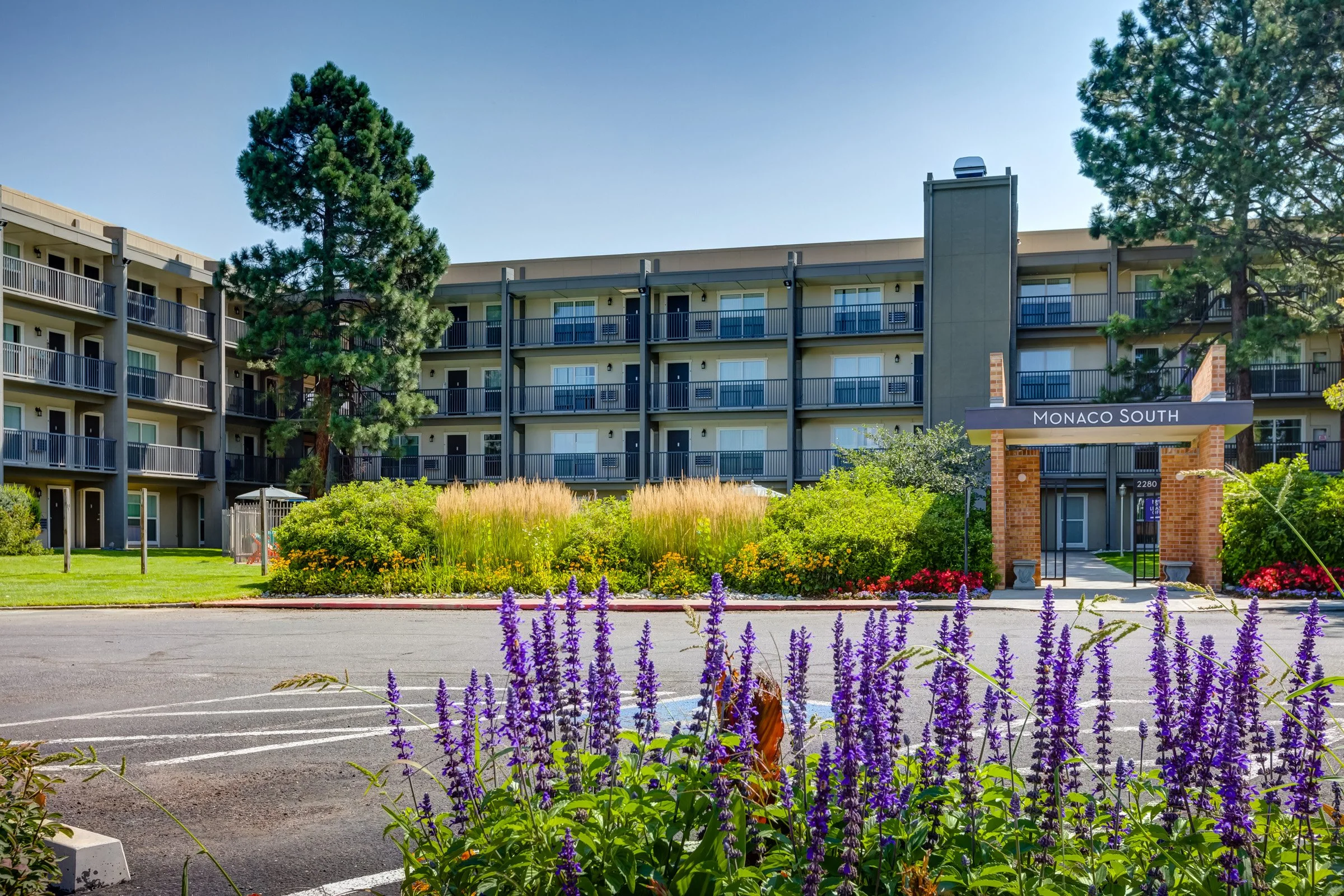 Exterior view of a multi-story apartment building called Monaco South, with balconies, surrounded by greenery, flowers, and tall trees, under a clear blue sky.