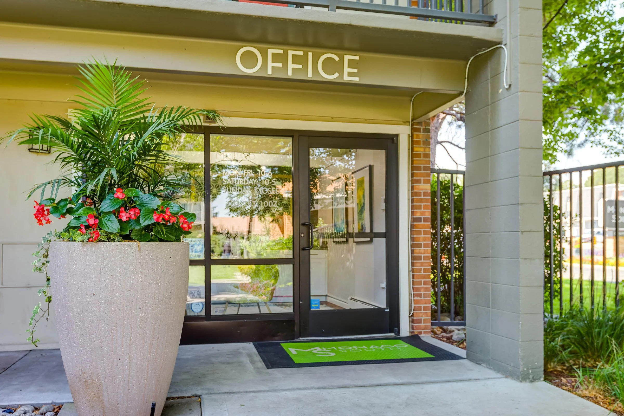 Office building entrance with glass door, large potted plant with green leaves and pink flowers, and a green Monash University welcome mat in front.