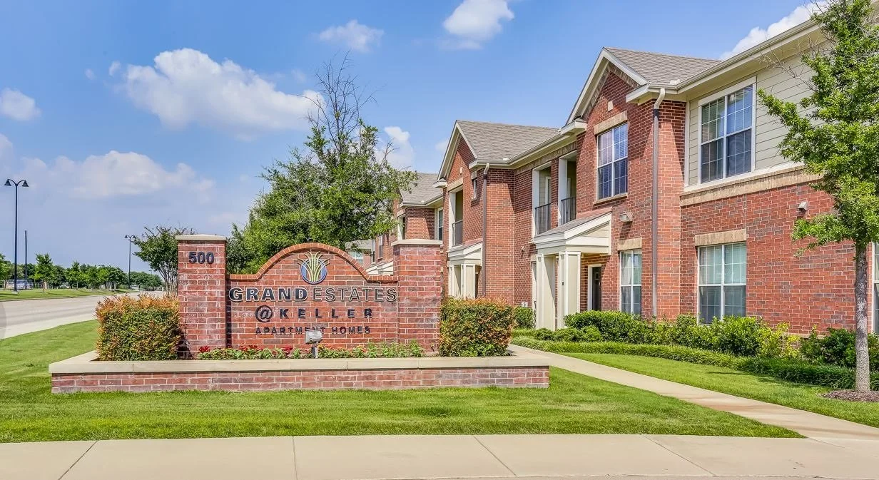 Photo of the entrance to Grand Estates at Keller apartment homes in a suburban neighborhood with brick buildings, green lawns, trees, and a sidewalk under a partly cloudy sky.