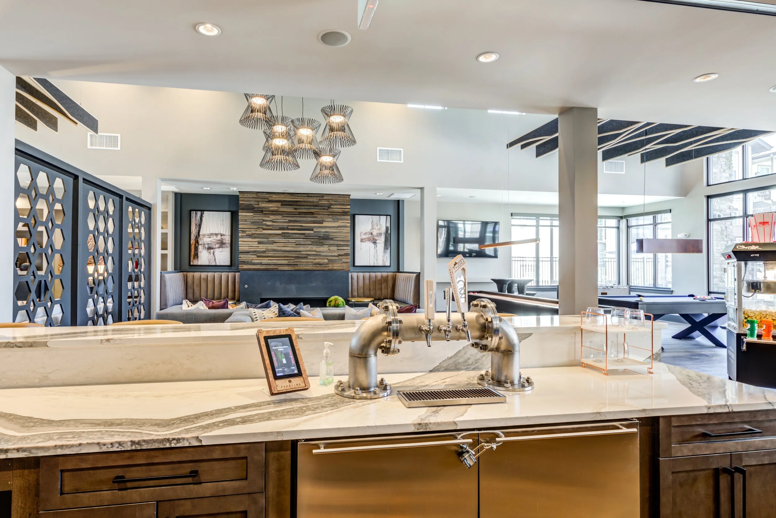 Modern indoor lounge with a bar counter in the foreground, featuring a marble countertop and stainless steel tap handles. The background shows comfortable seating with pillows, artwork on the walls, and large windows letting in natural light.