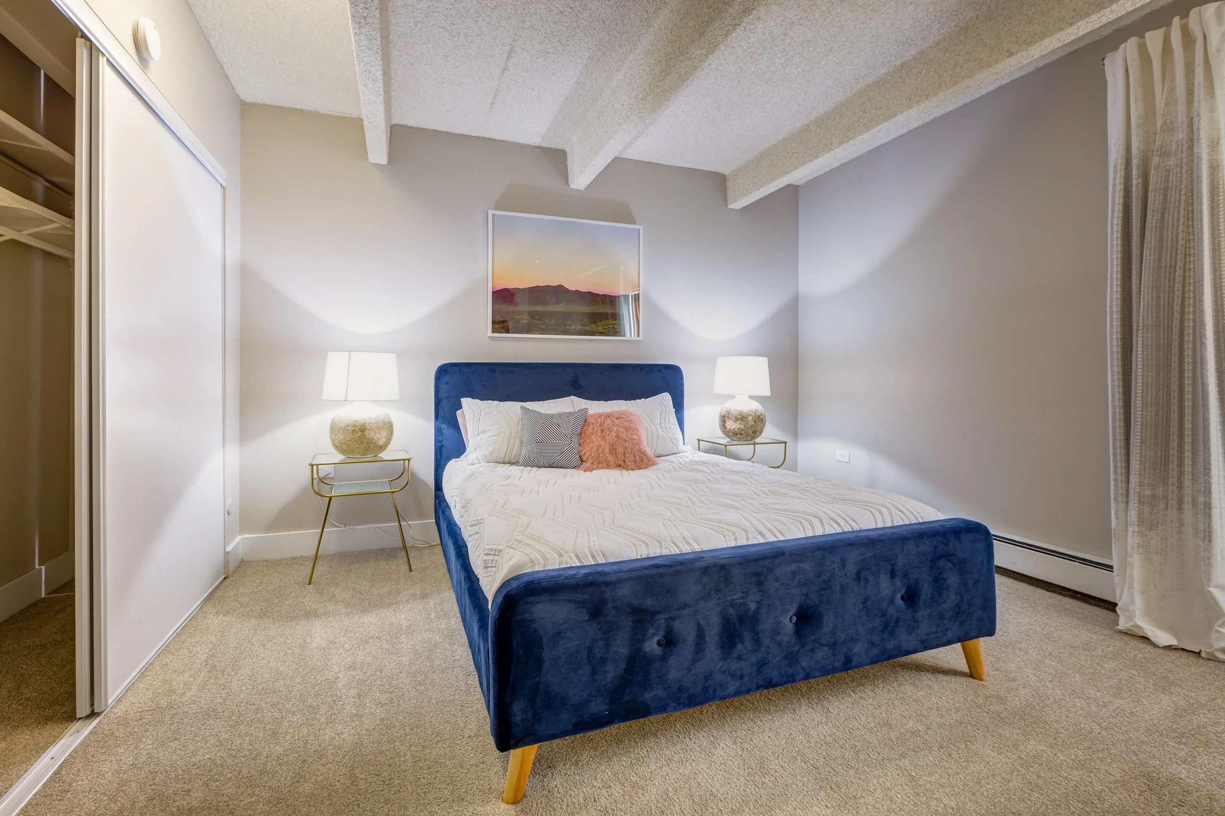 Cozy bedroom with a blue velvet bed, white bedding, two matching nightstands with lamps, wall art above the bed, and beige carpeted floor.