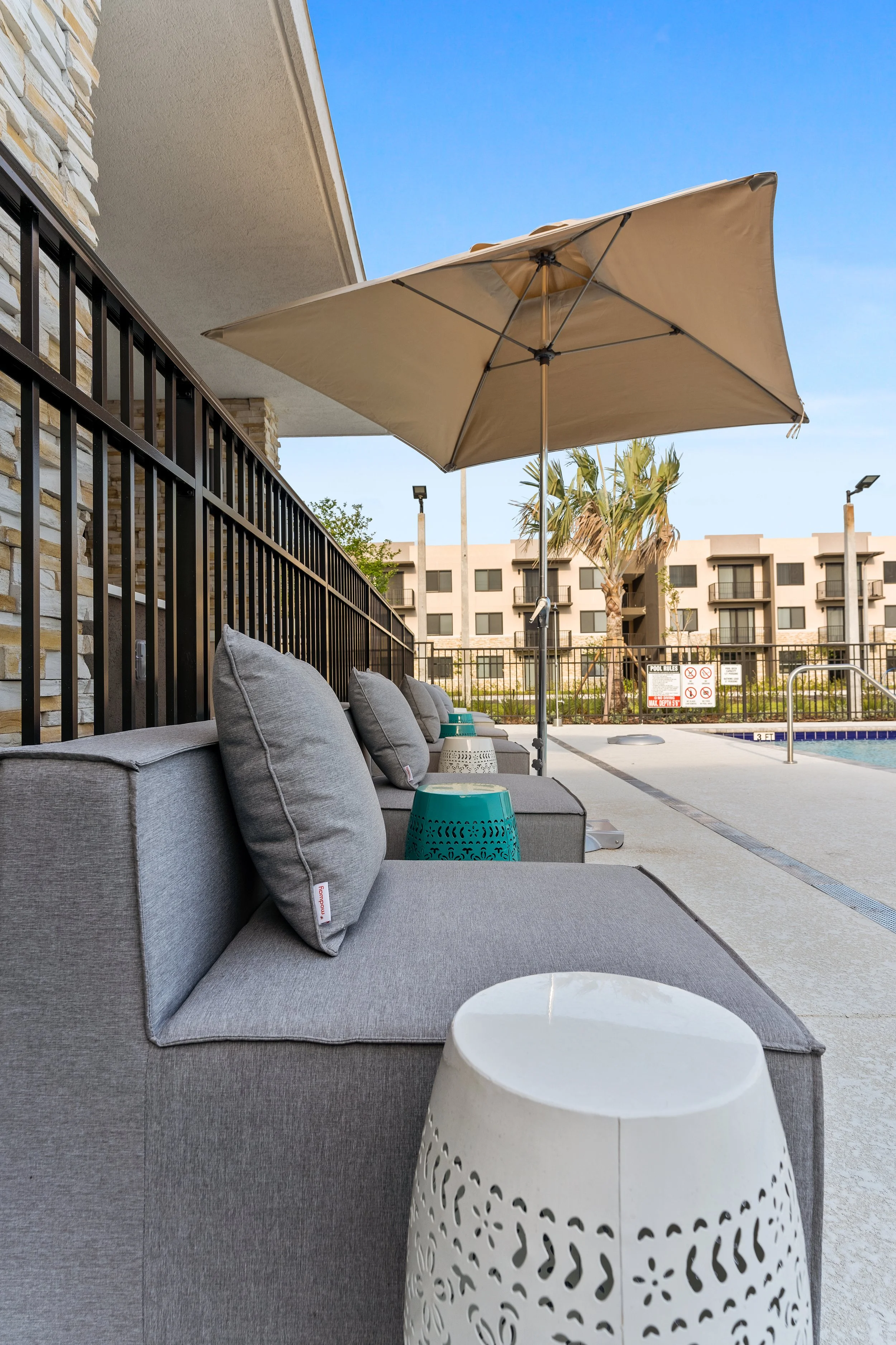 Row of outdoor lounge chairs with gray cushions and pillows, teal and white side tables, large beige umbrella, near swimming pool at a hotel or apartment complex, with palm trees and modern buildings in the background.