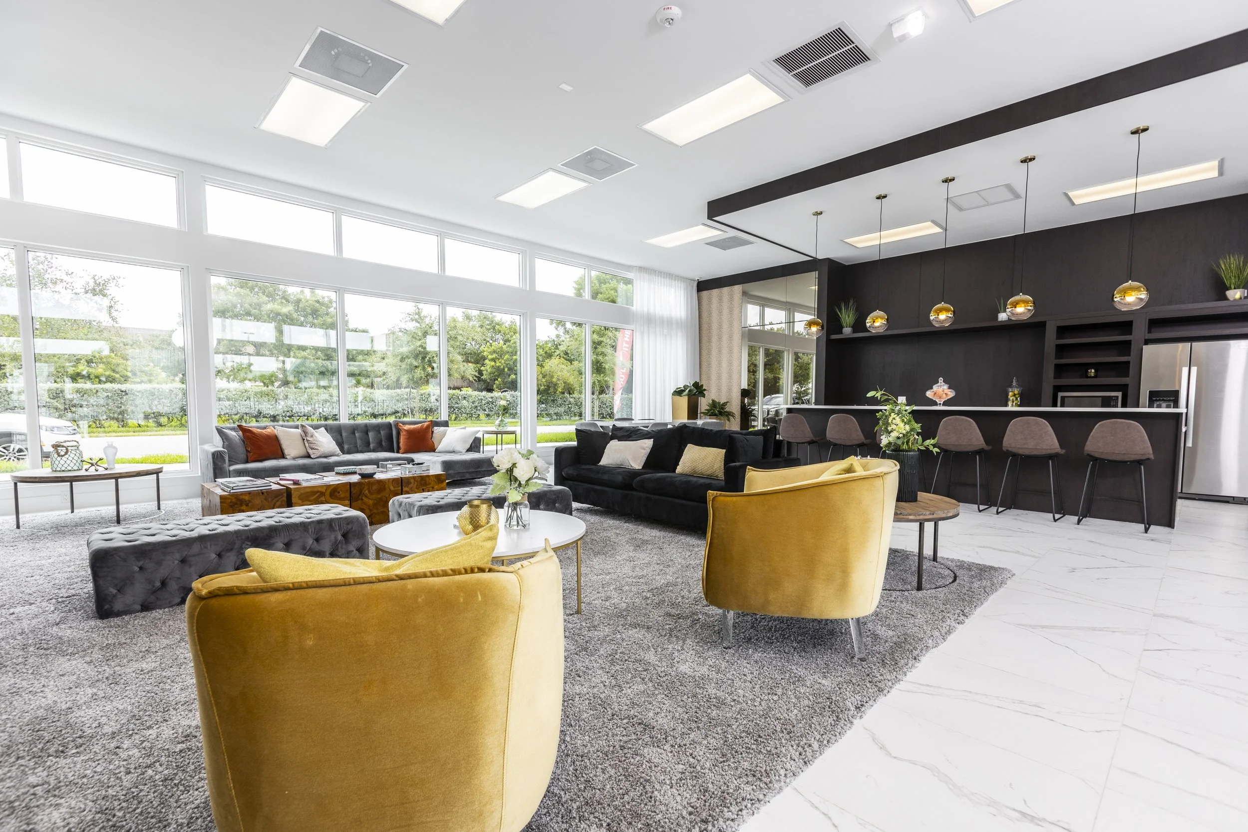 Modern lounge area with yellow velvet armchairs, black and gray sofas, white coffee tables, large windows with natural light, bar with pendant lights and seating, surrounded by greenery outside.