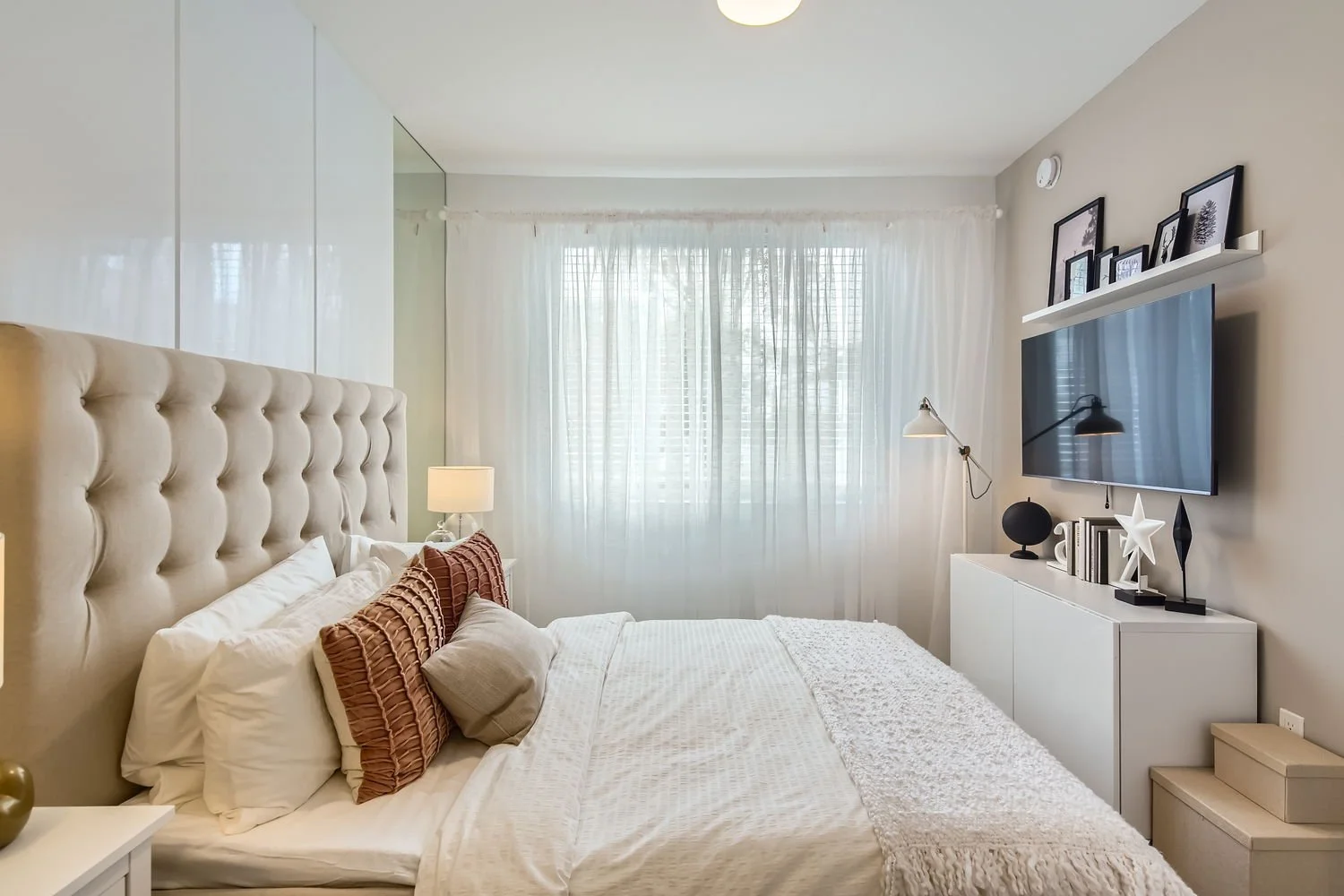 Modern bedroom with beige upholstered headboard, white bedding, decorative pillows, bedside lamp, window with sheer curtains, wall-mounted TV, and white console with framed photos and decorative objects.