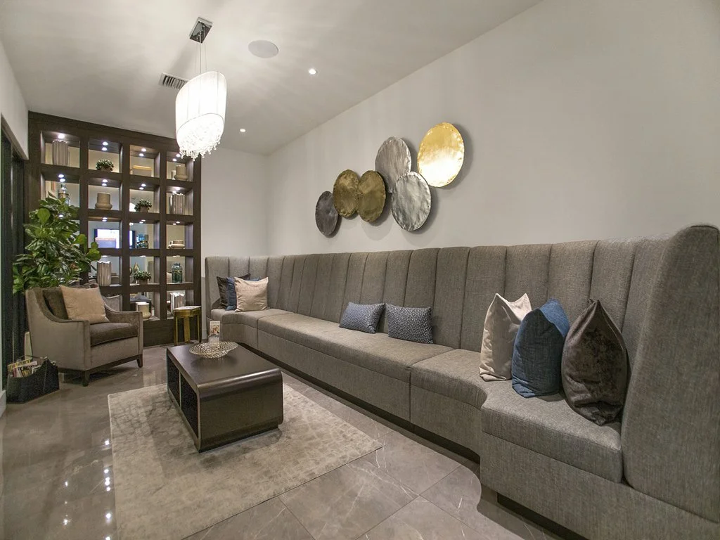 Living room with a large gray sectional sofa, decorative pillows, a coffee table, a single armchair, a shelving unit with plants and ornaments, artwork on the wall, and a hanging ceiling light fixture.